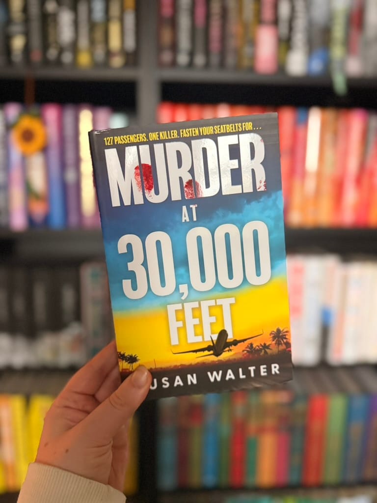 Hand holding the book Murder at 30,000 Feet by Susan Walter in front of colorful blurred bookshelves.