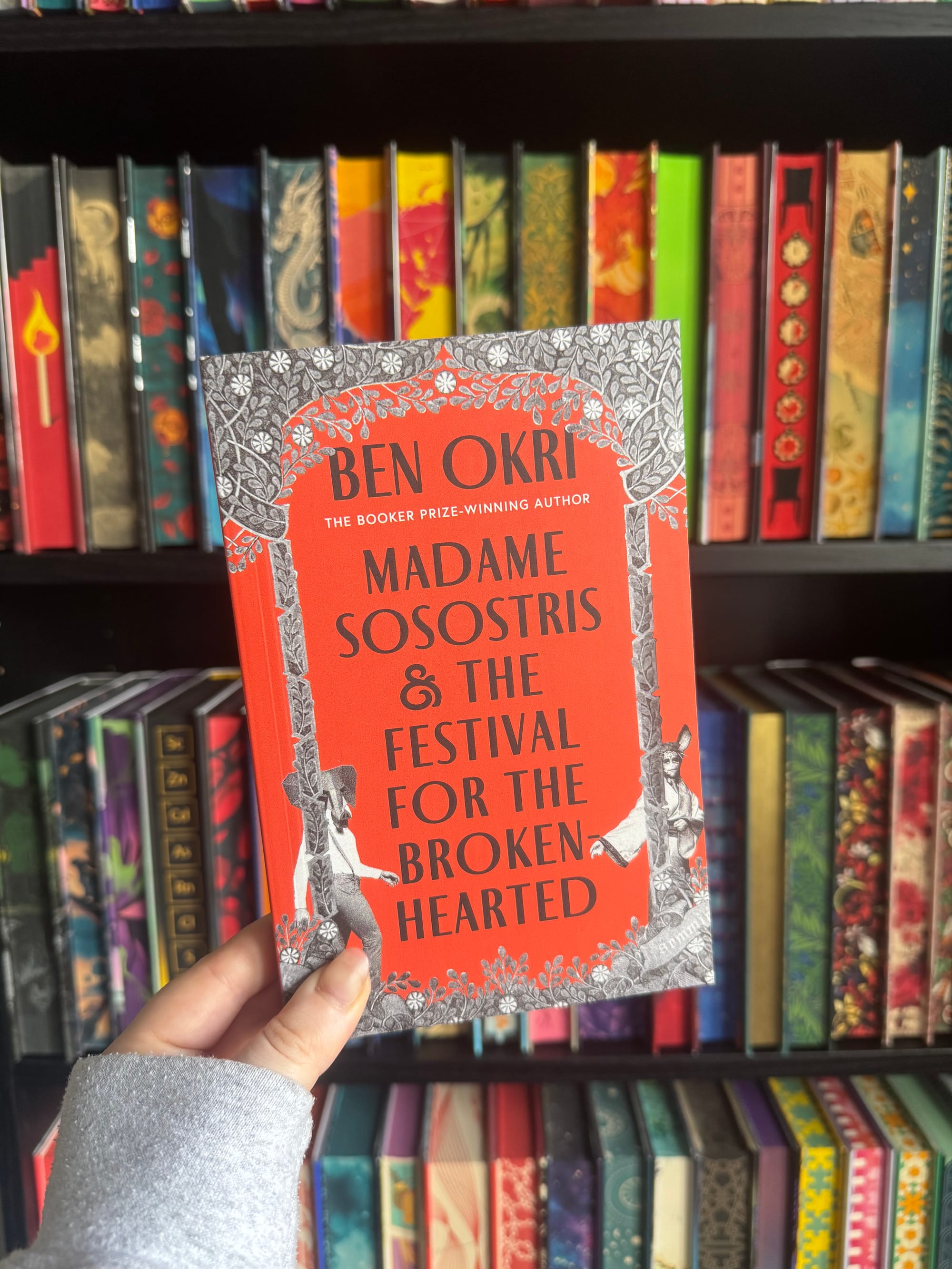 A hand holds a proof copy of Madame Sosostris & The Festival for the Brokenhearted by Ben Okri in front of softly blurred rainbow bookshelves. The striking cover features a vivid red centre framed by intricate black and grey floral illustrations, with small theatrical figures positioned around the border. The bold typography and ornate design hint at the book's magical, literary atmosphere.