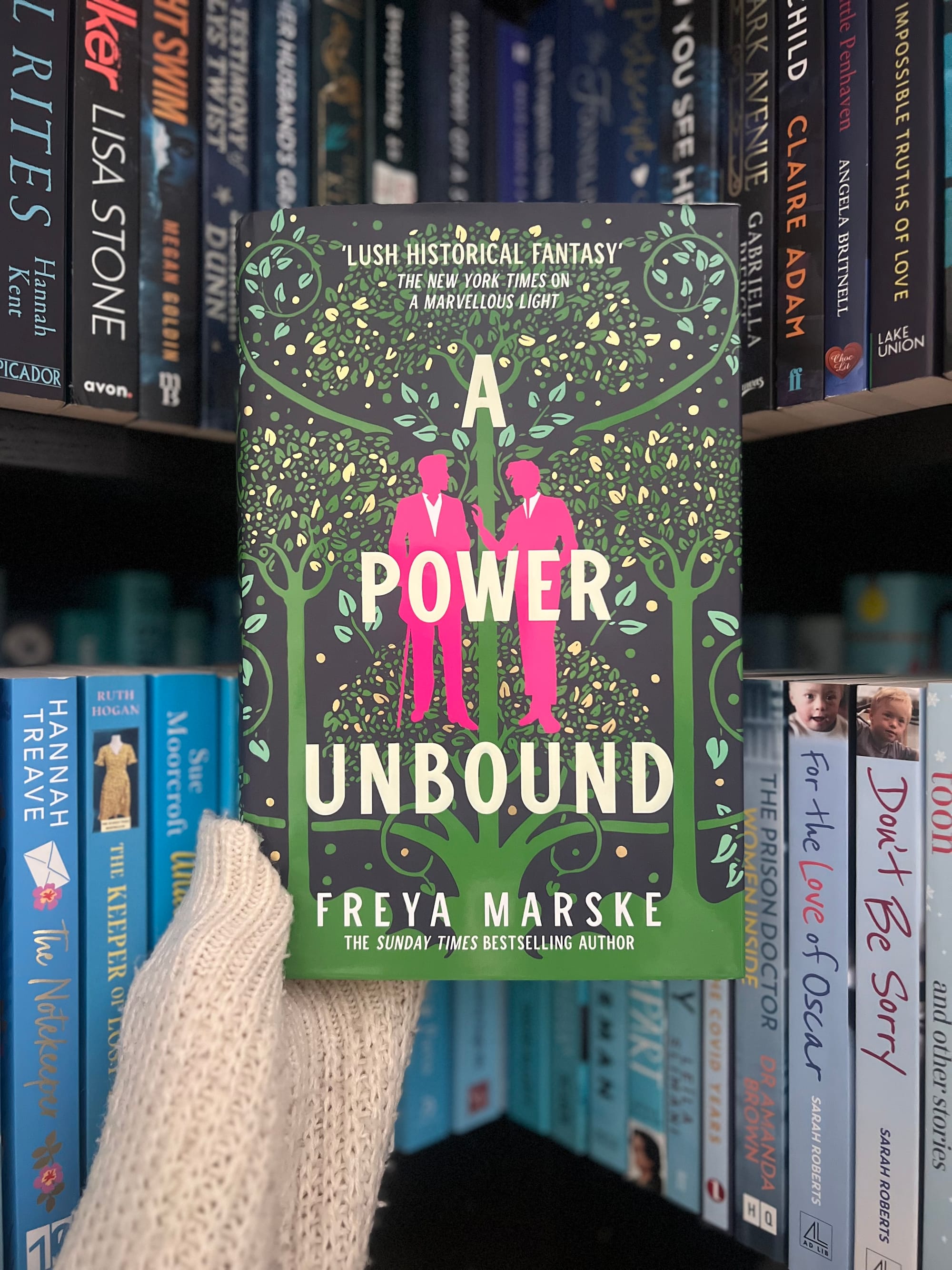 A gloved hand holds a book titled "A Power Unbound" by Freya Marske. The cover features green and pink designs. Bookshelves are in the background.