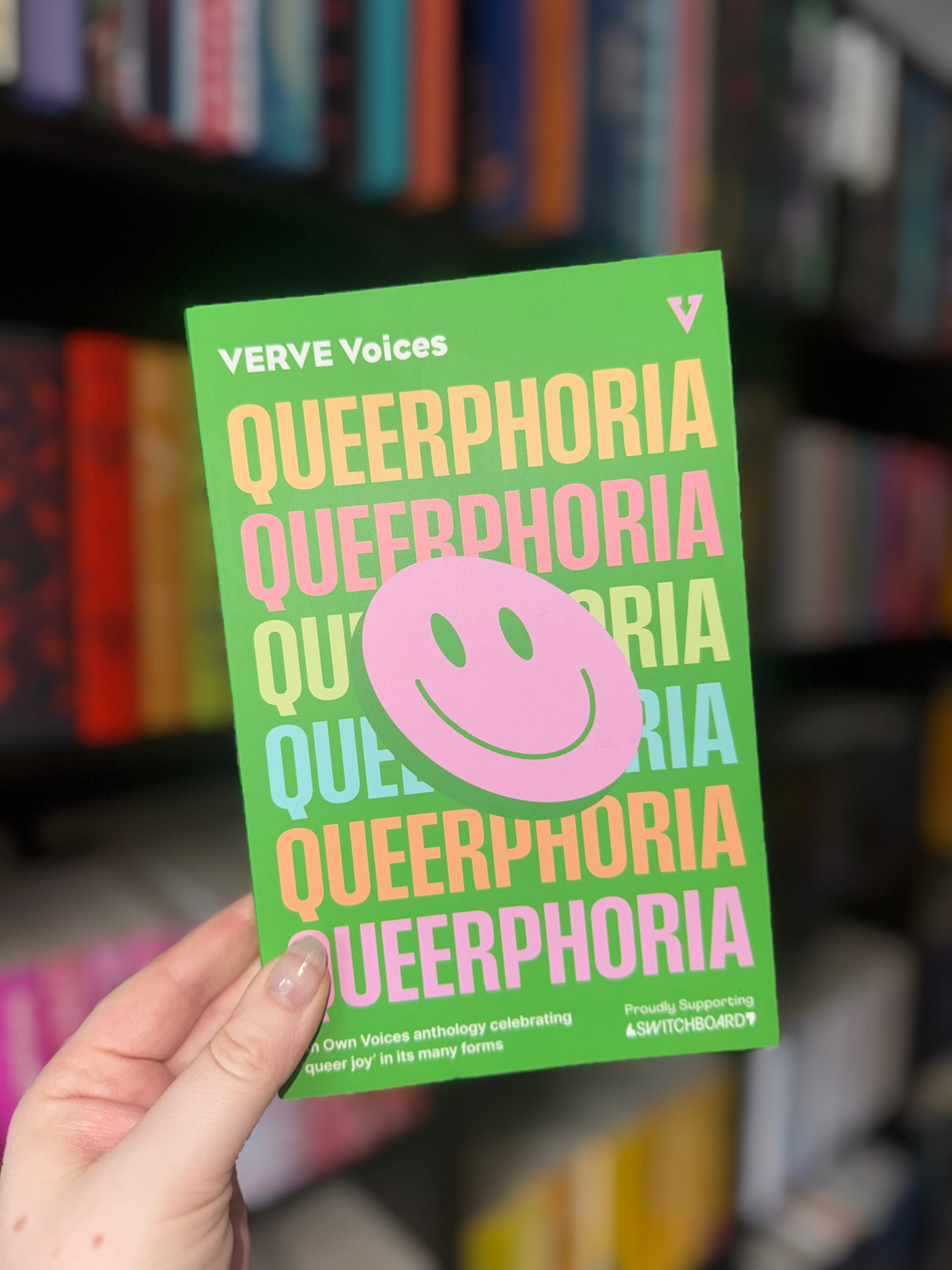 Hand holds a book titled "QUEERPHORIA" in bold colors with a pink smiley sticker. Background shows blurred books on shelves. Bright and lively mood.
