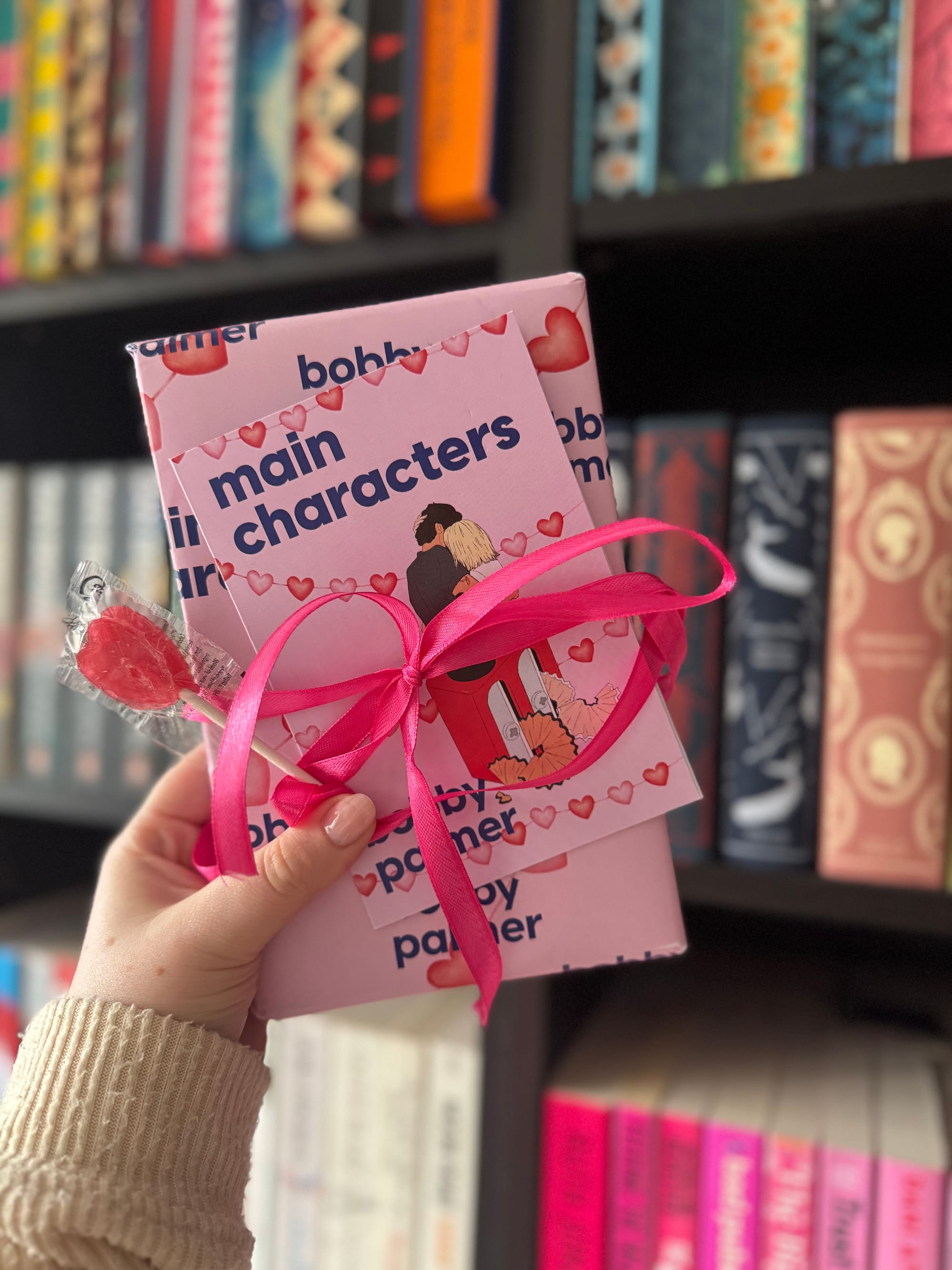 Hand holding wrapped books titled "main characters" with hearts, pink ribbon, and a heart lollipop, set against a bookshelf background.