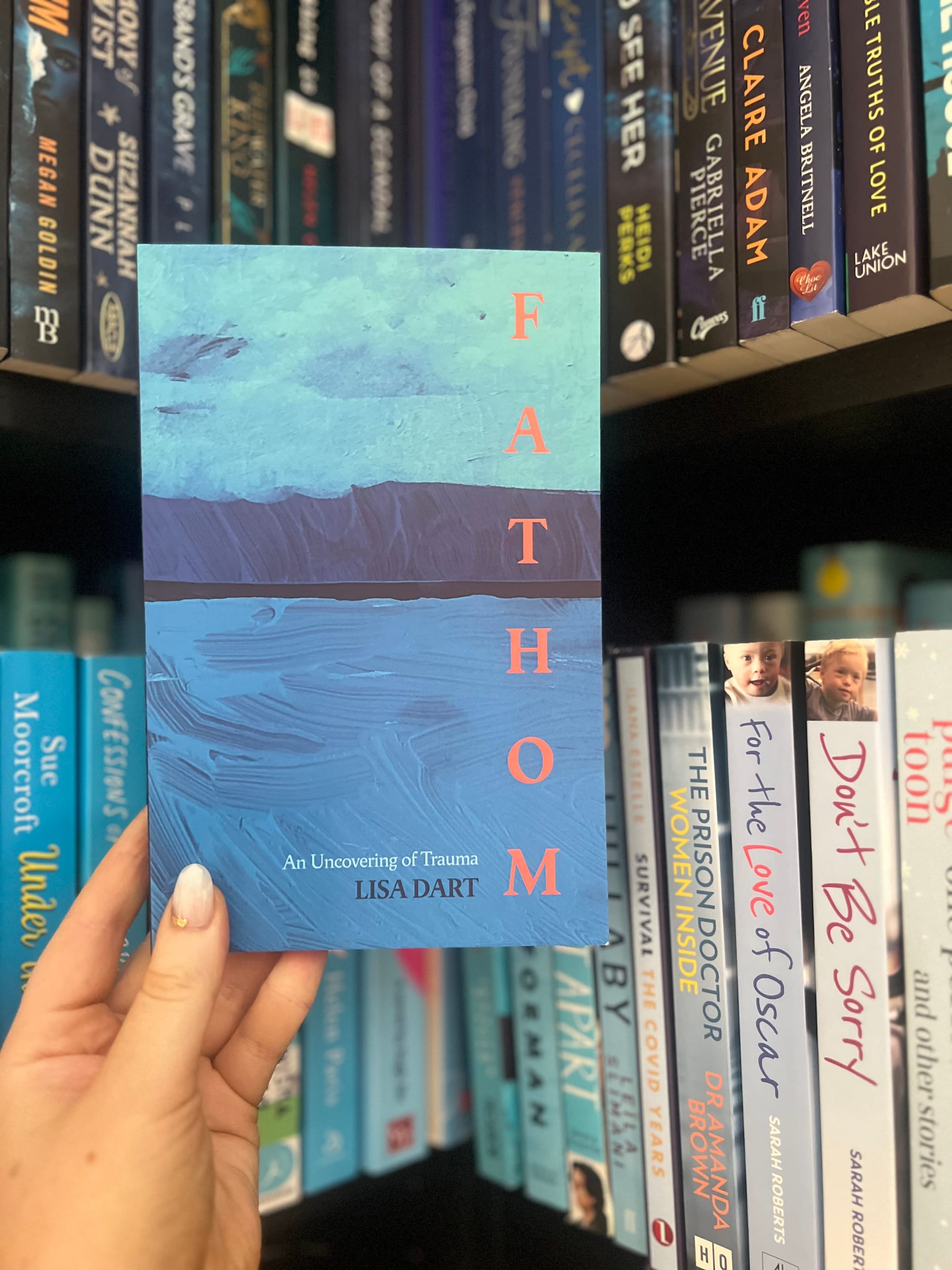 A hand holds the book "Fathom" by Lisa Dart, with a blue sea-themed cover, in front of a shelf of assorted books in a cozy bookstore.