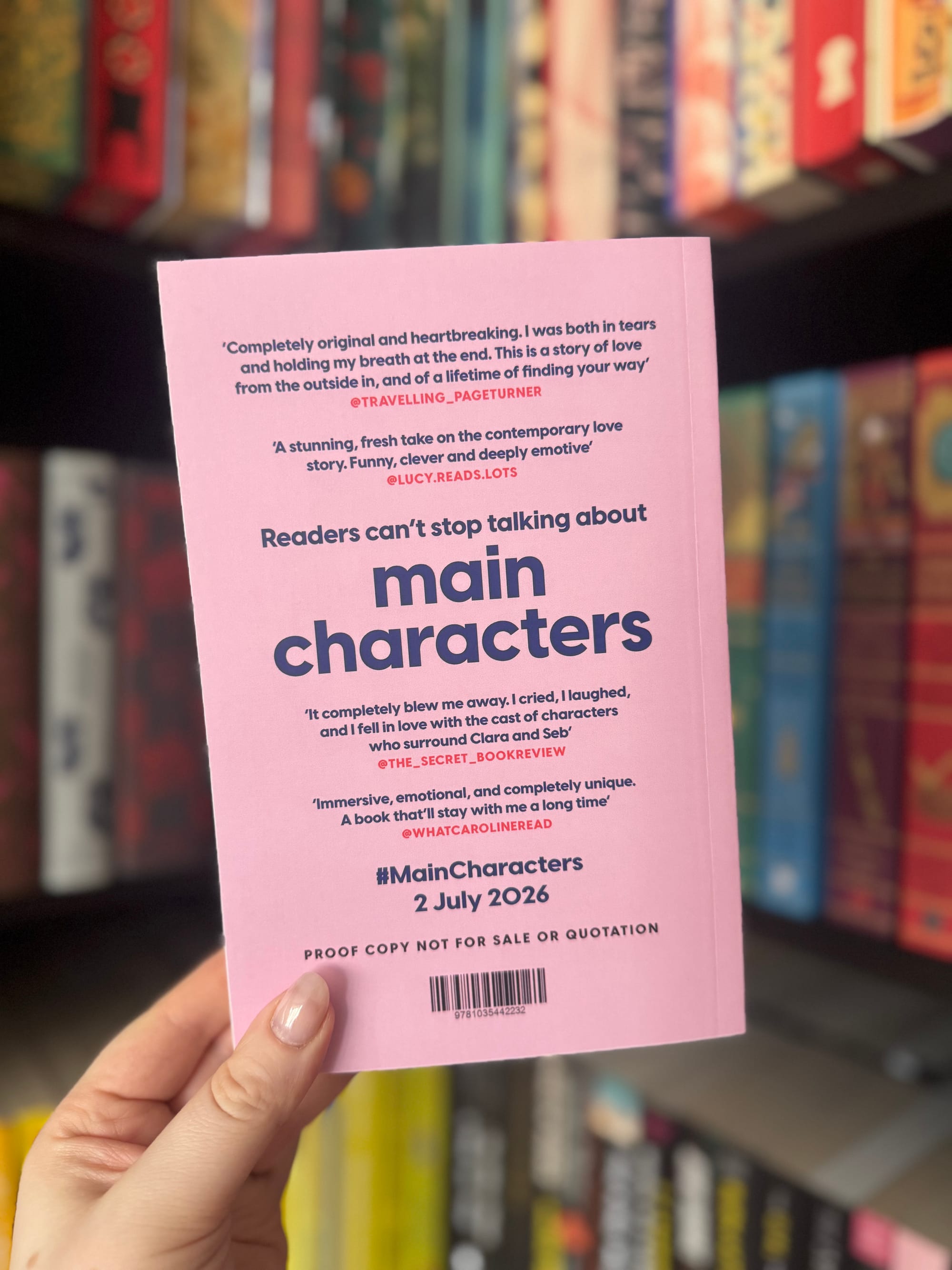 Back cover of the Main Characters proof copy showing early praise quotes and the publication date July 2026, held in front of a bookshelf.