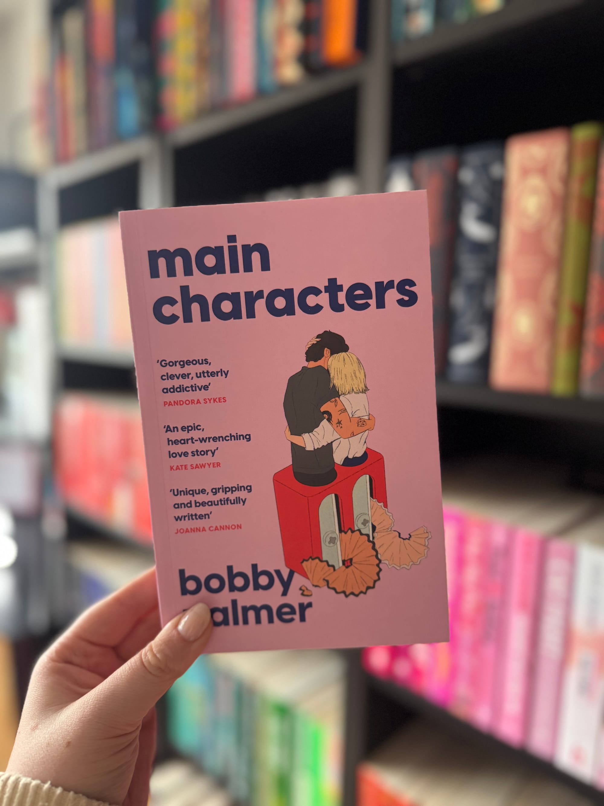 Close up of the pink proof cover of Main Characters by Bobby Palmer held in front of a blurred bookshelf filled with colourful books.