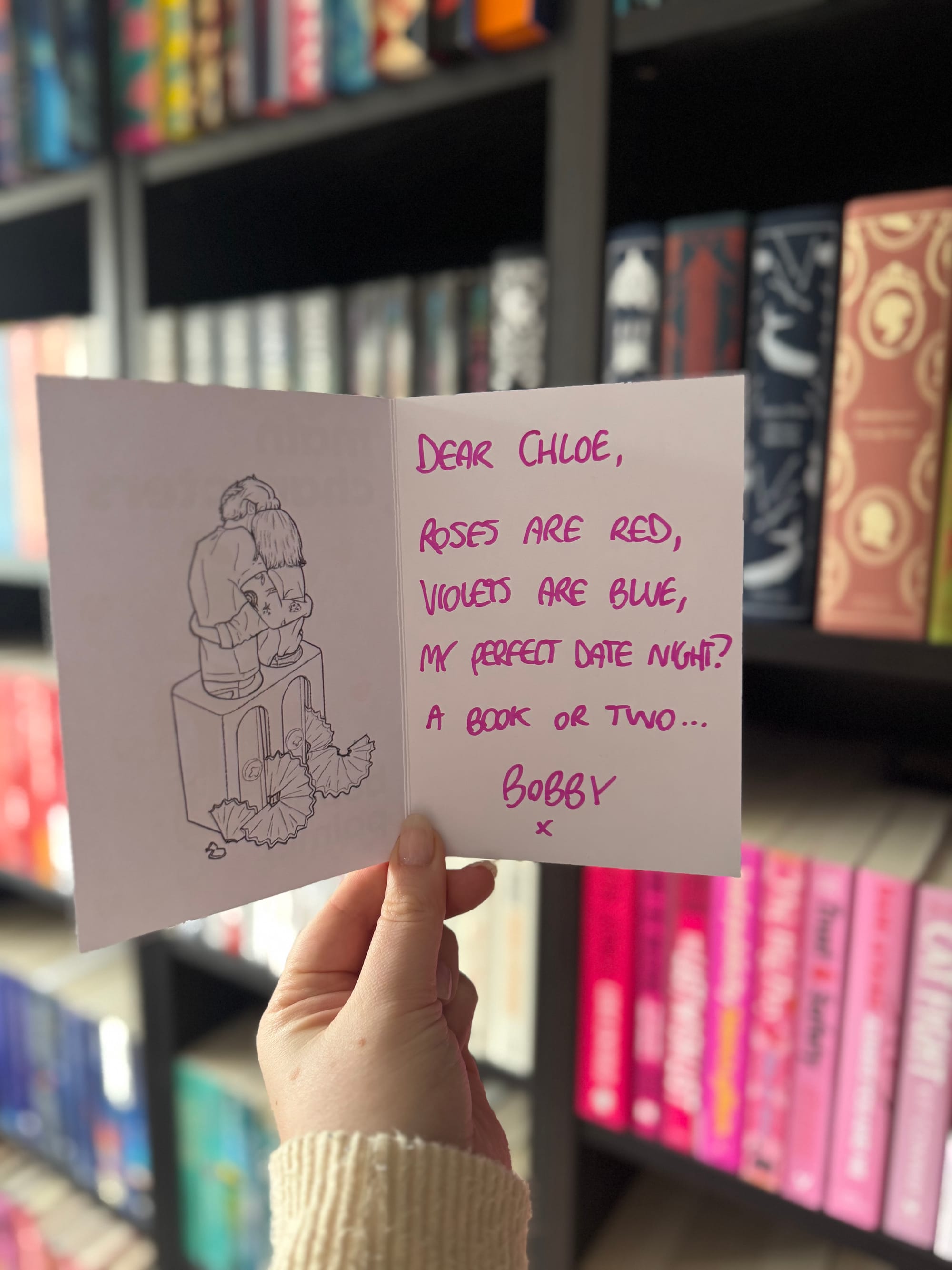 Open card inside the Main Characters proof copy showing a handwritten message that reads “Dear Chloe, Roses are red, violets are blue, my perfect date night? A book or two… Bobby” with a small illustration of a couple sitting together.