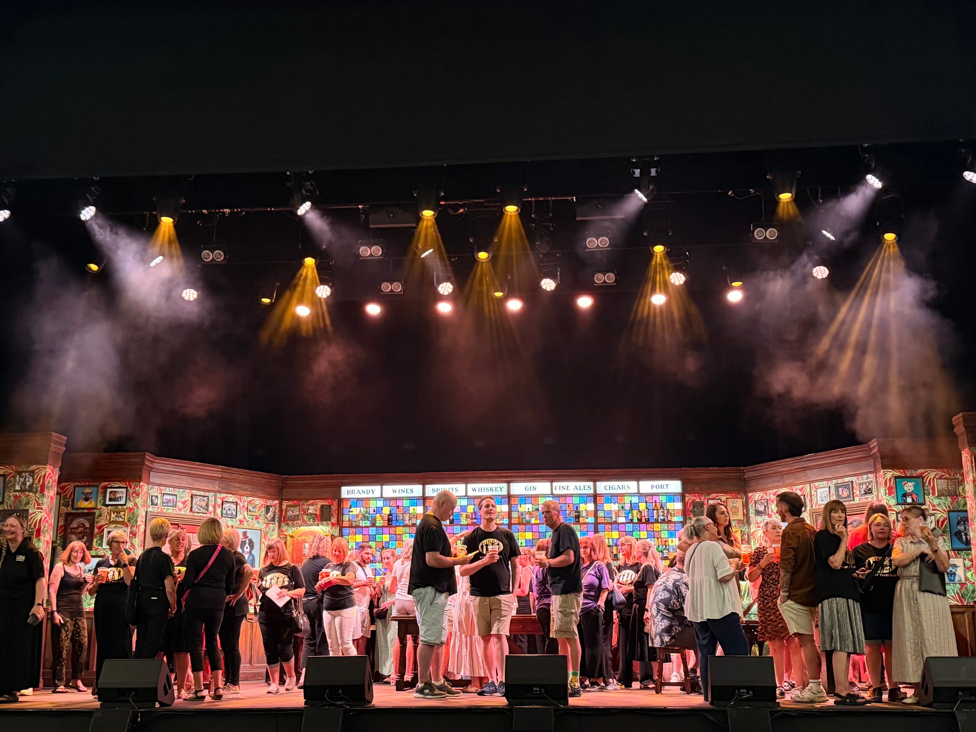 People mingle on a theater stage under warm spotlights, with a colorful bar backdrop labeled brandy, wines, spirits, whiskey, gin.