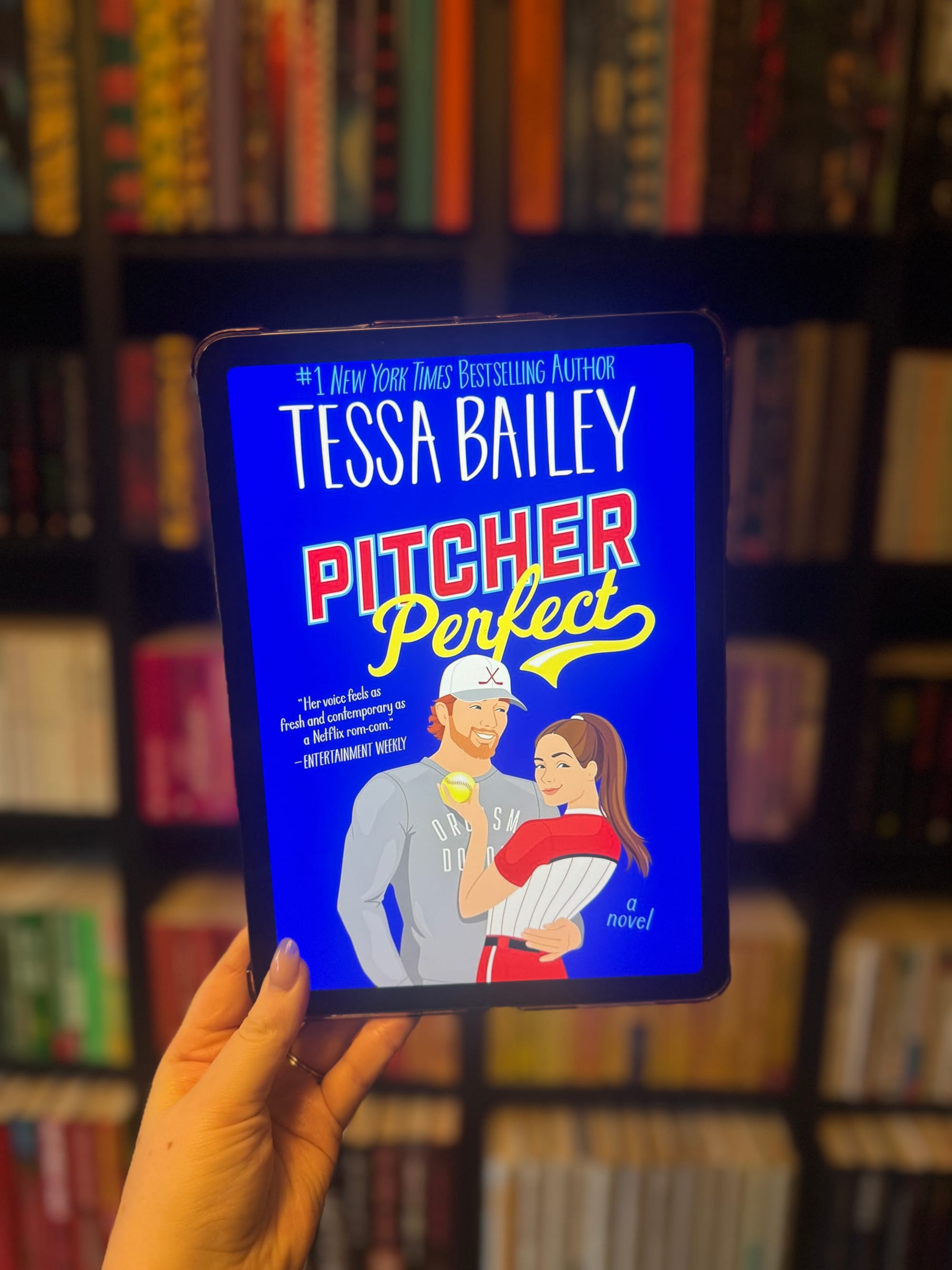 Hand holding a tablet displaying the book cover "Pitcher Perfect" by Tessa Bailey. Cartoon couple; baseball theme; bookshelves in the background.