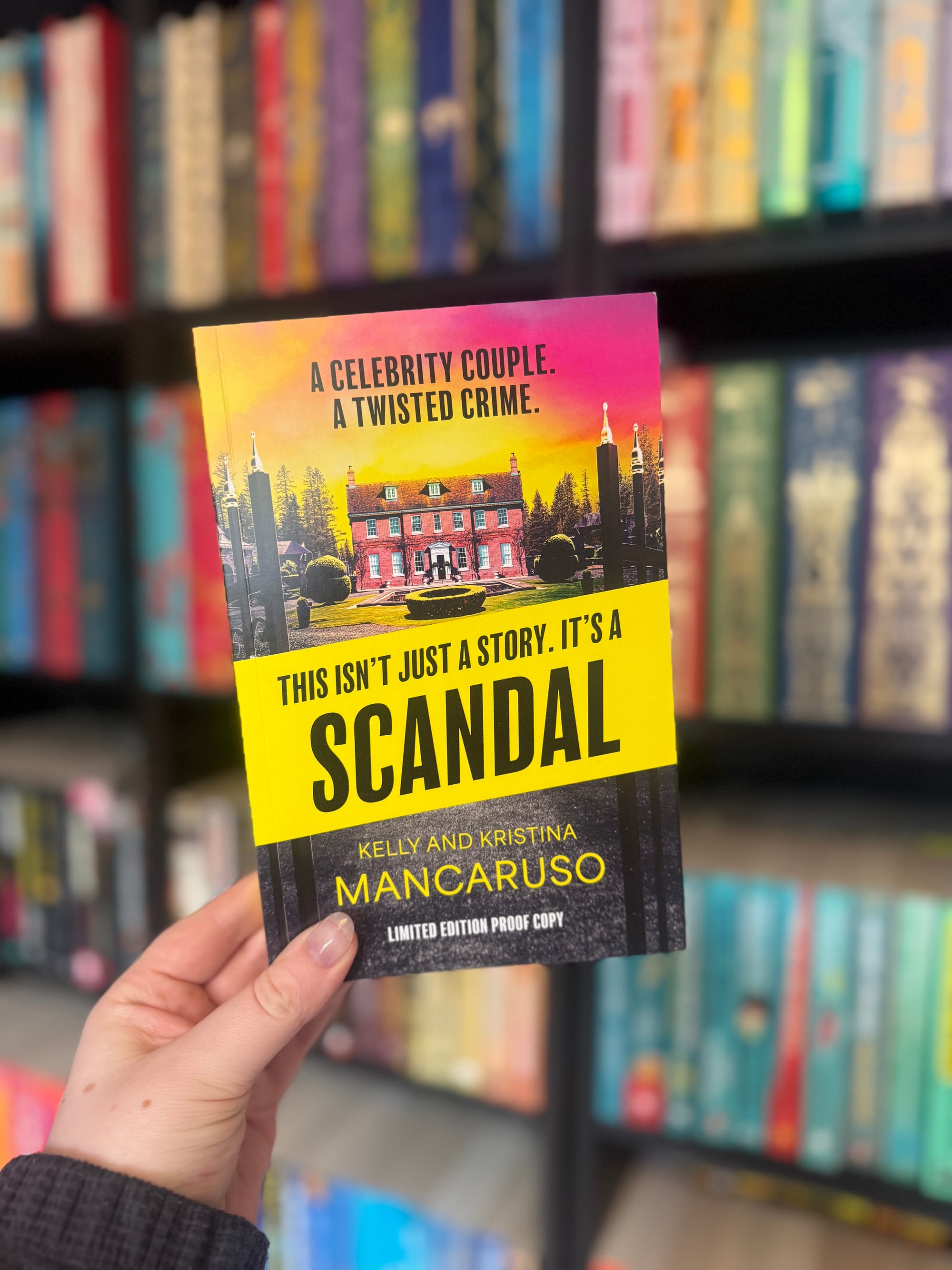Hand holding a book titled "Scandal" with a vibrant cover, in front of colorful blurred bookshelves. Text: "A celebrity couple. A twisted crime."