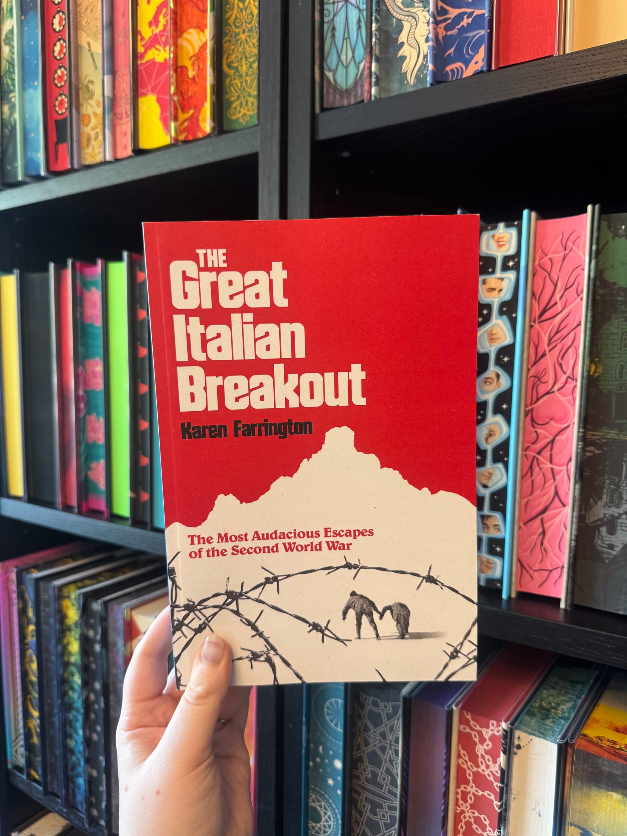 Hand holding the red book The Great Italian Breakout by Karen Farrington in front of colorful bookshelves.