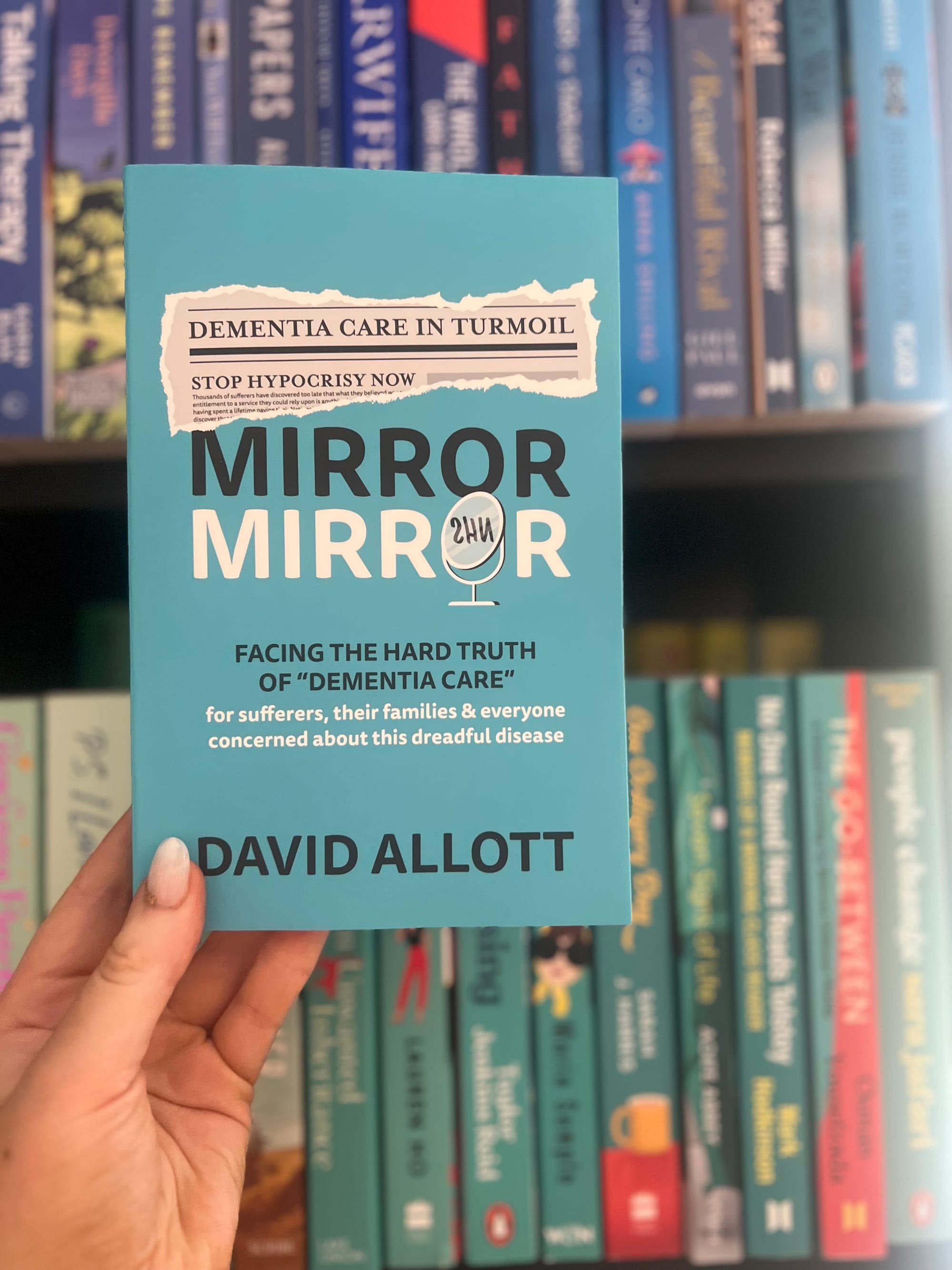 Hand holding a blue book titled "Mirror Mirror" by David Allott, about dementia care. Bookshelf with various colorful books in the background.