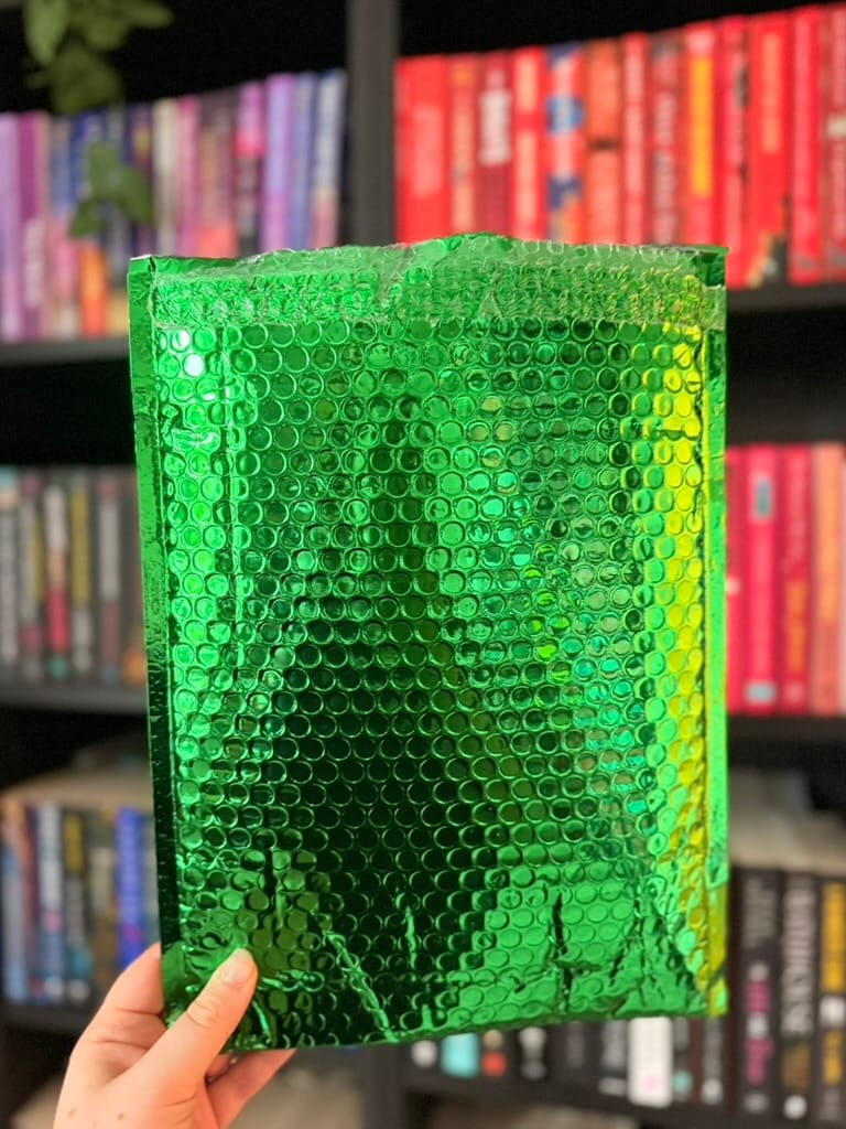 A hand holds a shiny green padded envelope in front of blurred, colour-coordinated bookshelves.