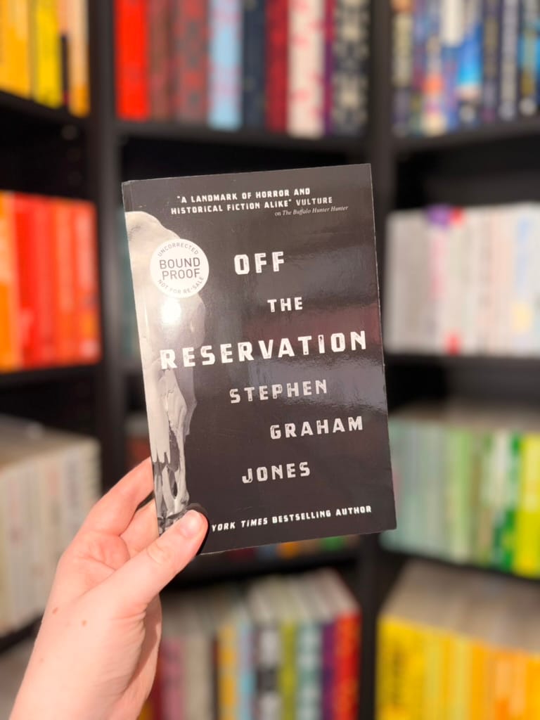 A hand holds a bound proof copy of Off the Reservation by Stephen Graham Jones in front of softly blurred, colour-organised bookshelves. The dark cover features bold white lettering and a partially visible skull illustration, with a circular ‘Uncorrected Bound Proof’ label.