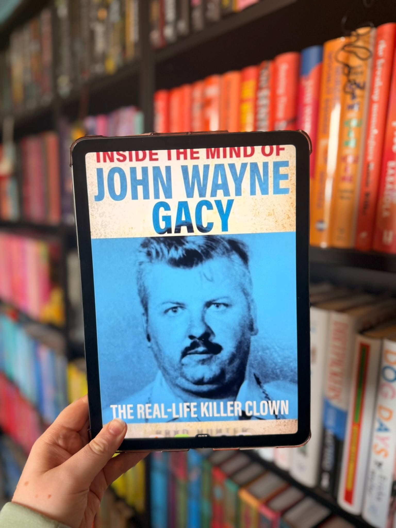 A hand holds an e-reader displaying the cover of Inside the Mind of John Wayne Gacy: The Real-Life Killer Clown by Christopher Berry-Dee in front of softly blurred rainbow bookshelves. The cover features a blue-tinted mugshot of John Wayne Gacy beneath bold blue and red title text, with the subtitle highlighting the book's focus on the infamous serial killer. The stark, documentary-style design reflects the book's true crime subject matter.