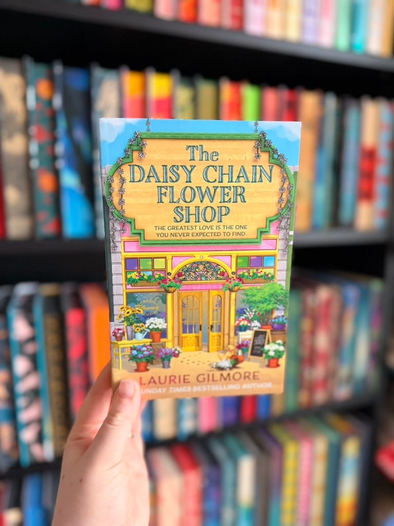 A hand holds an e-reader displaying the cover of The Daisy Chain Flower Shop by Laurie Gilmore in front of softly blurred rainbow bookshelves. The colourful cover features a charming flower shop with a pastel pink façade, bright yellow doors, overflowing window boxes, potted flowers, and a small dog sitting outside. The cheerful illustration and vibrant blooms reflect the book's cosy, romantic atmosphere.