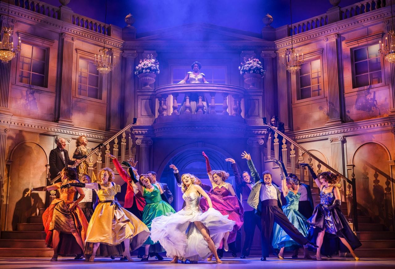 The cast of High Society performing on an elaborate mansion set, dressed in colourful formal costumes beneath chandeliers and dramatic stage lighting.