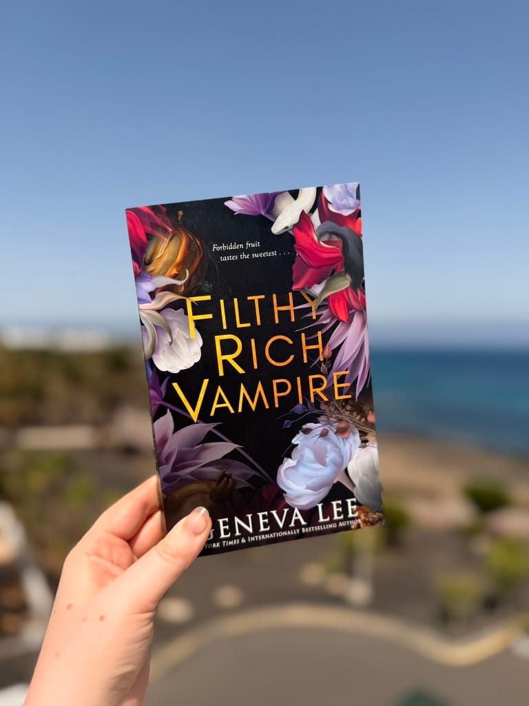 A hand holds a copy of Filthy Rich Vampire by Geneva Lee outdoors against a softly blurred coastal backdrop. The dark book cover features vivid red, purple and white flowers with bold gold lettering, contrasting against the bright blue sky and sea.