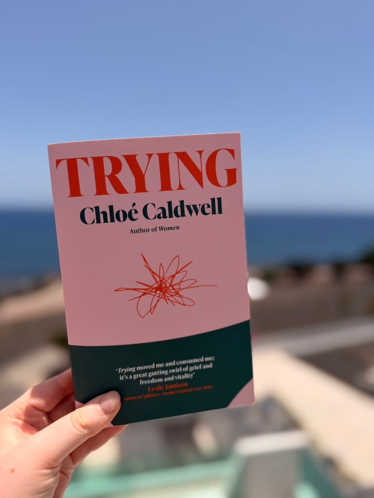 A hand holds a copy of Trying by Chloé Caldwell outdoors against a softly blurred coastal backdrop. The book has a pale pink cover with bold red and dark green typography, while the blue sky and sea create a bright, summery background.