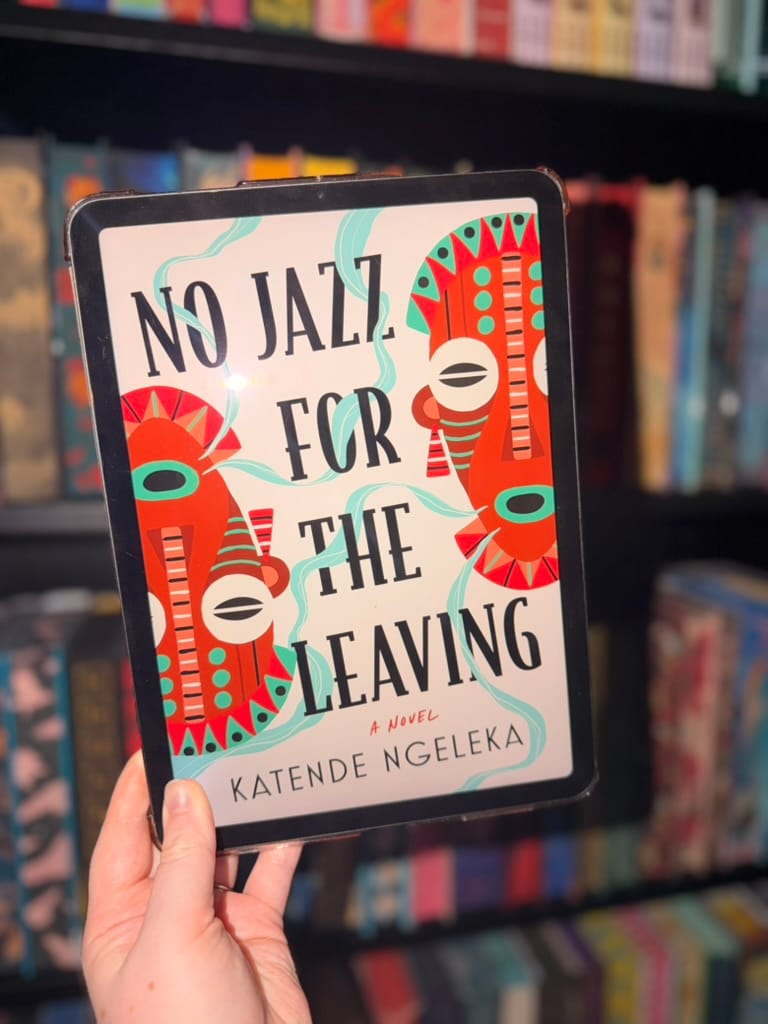 A hand holds an e-reader displaying the cover of No Jazz for the Leaving by Katende Ngeleka in front of softly blurred rainbow bookshelves. The vibrant cover features bold black typography surrounded by striking red, turquoise and black African-inspired masks, connected by flowing turquoise lines across a white background. The colourful, graphic design creates a distinctive and eye-catching look that reflects the book's rich cultural themes.