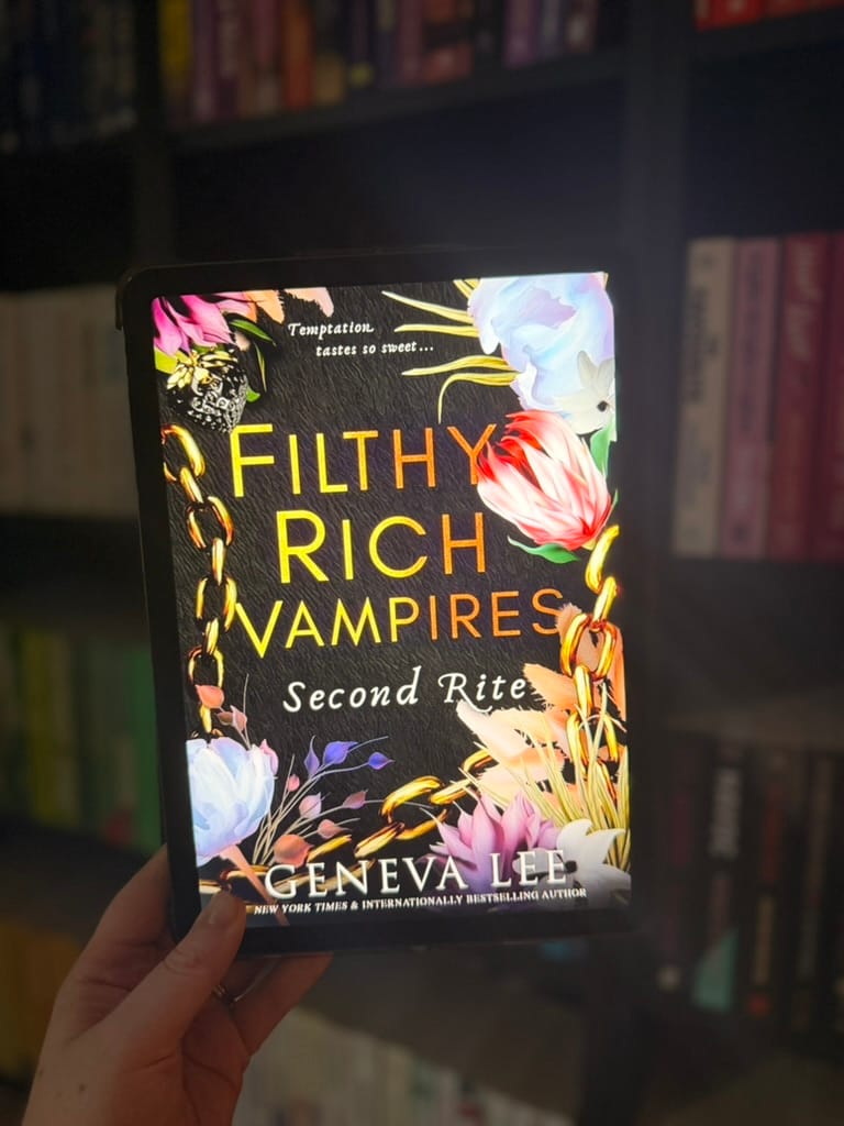 A hand holds a tablet displaying the cover of Second Rite, the second Filthy Rich Vampires book by Geneva Lee. The black cover features colourful flowers, gold chains and bold gold lettering, photographed against softly blurred bookshelves filled with colourful books.