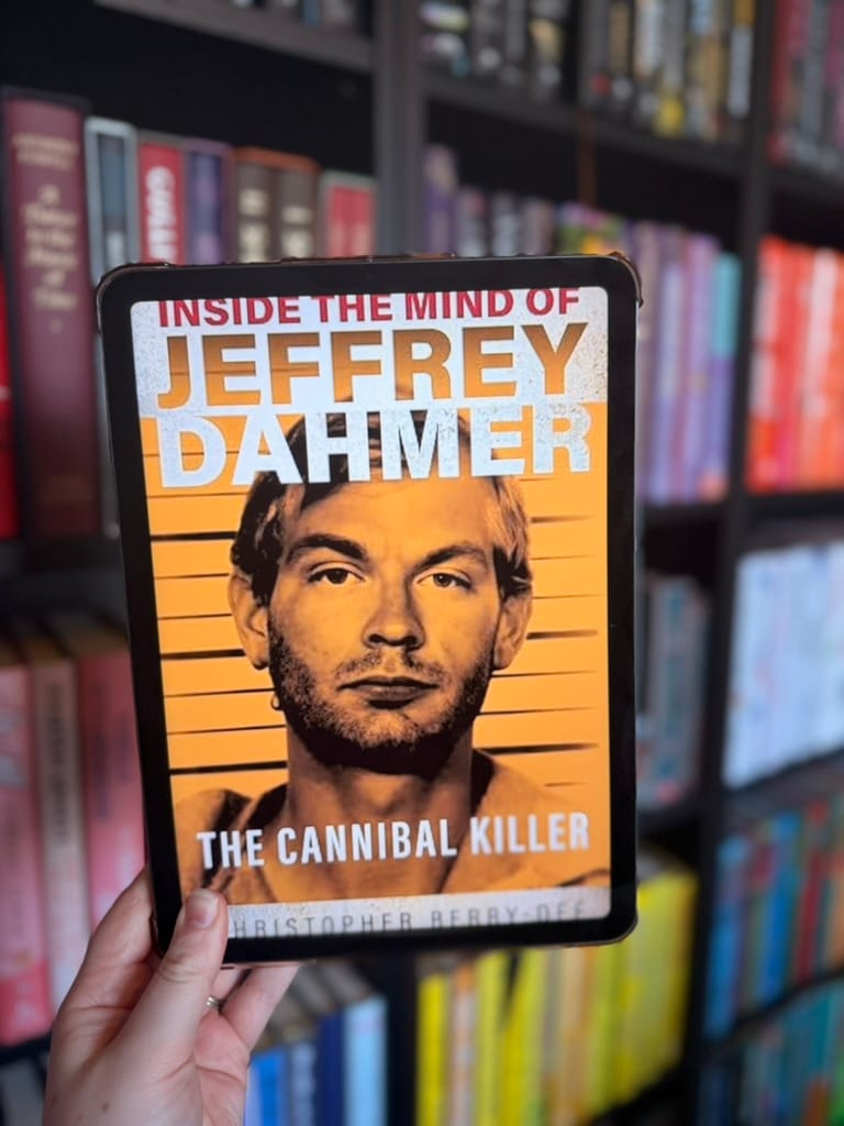 A hand holds an e-reader displaying the cover of Inside the Mind of Jeffrey Dahmer: The Cannibal Killer by Christopher Berry-Dee in front of softly blurred rainbow bookshelves. The cover features a sepia-toned mugshot of Jeffrey Dahmer against a police lineup background, with bold white and orange text identifying the subject. The stark, documentary-style design reflects the book's true crime focus.