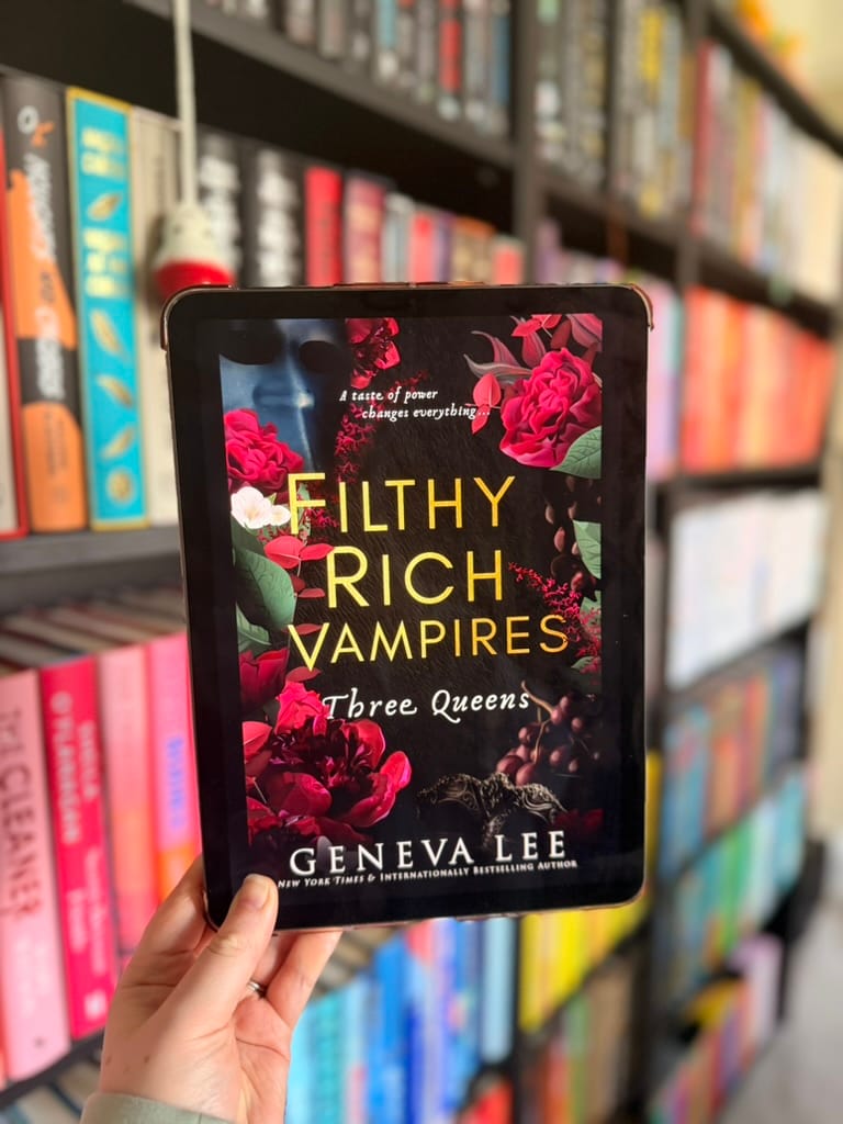 A hand holds a tablet displaying the cover of Three Queens from Geneva Lee’s Filthy Rich Vampires series. The dark cover features vivid red roses, gold lettering and ornate details, photographed against softly blurred, colour-organised bookshelves.