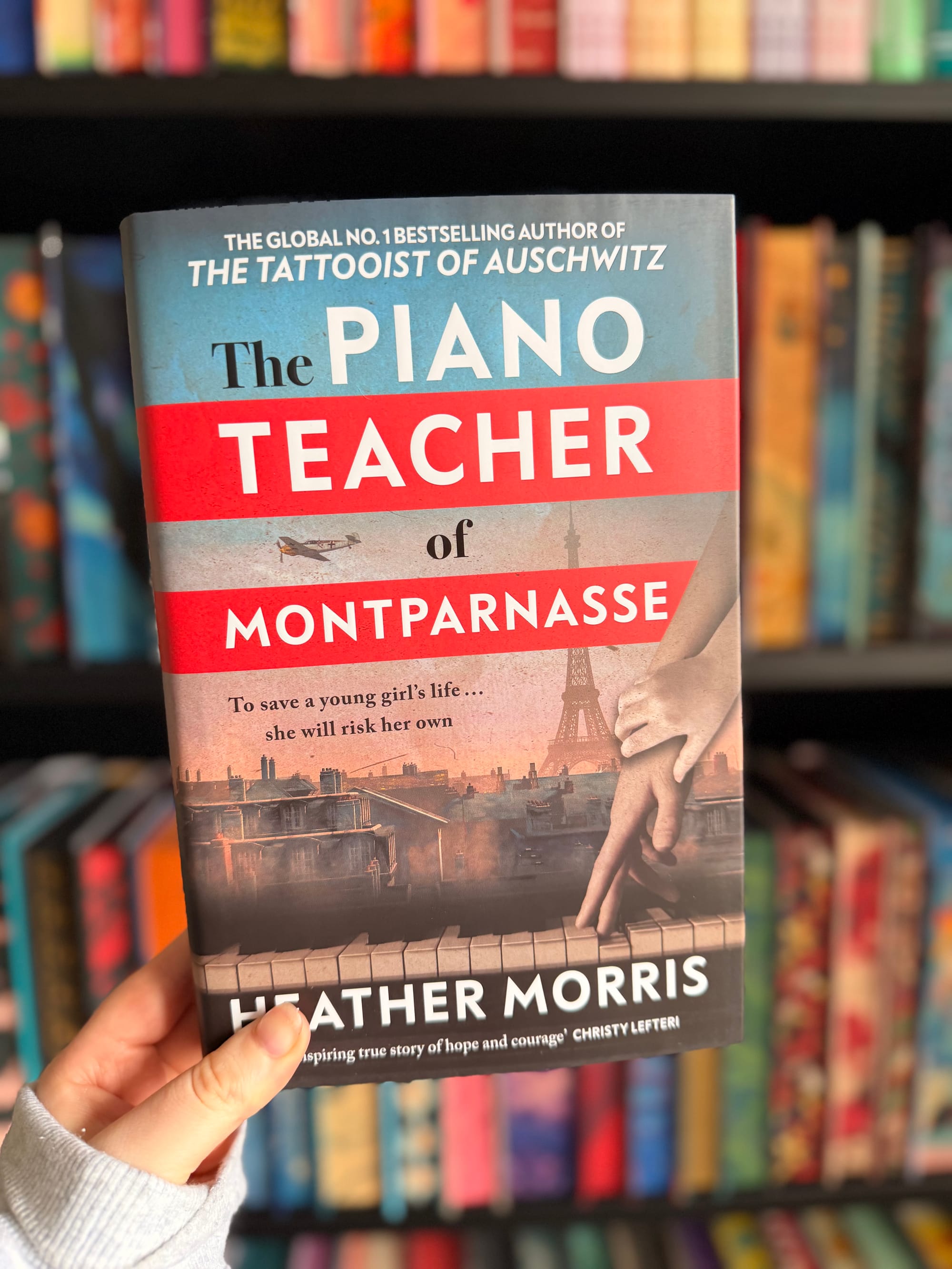 A hand holding a copy of The Piano Teacher of Montparnasse by Heather Morris in front of a colourful, softly blurred bookshelf filled with books.