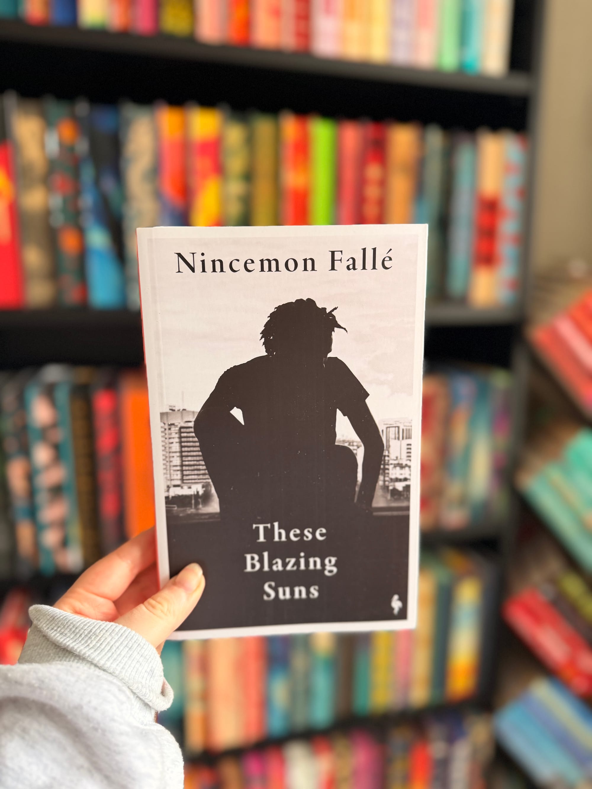 A hand holding a copy of These Blazing Suns by Nincemon Fallé in front of a colourful, softly blurred bookshelf filled with books.