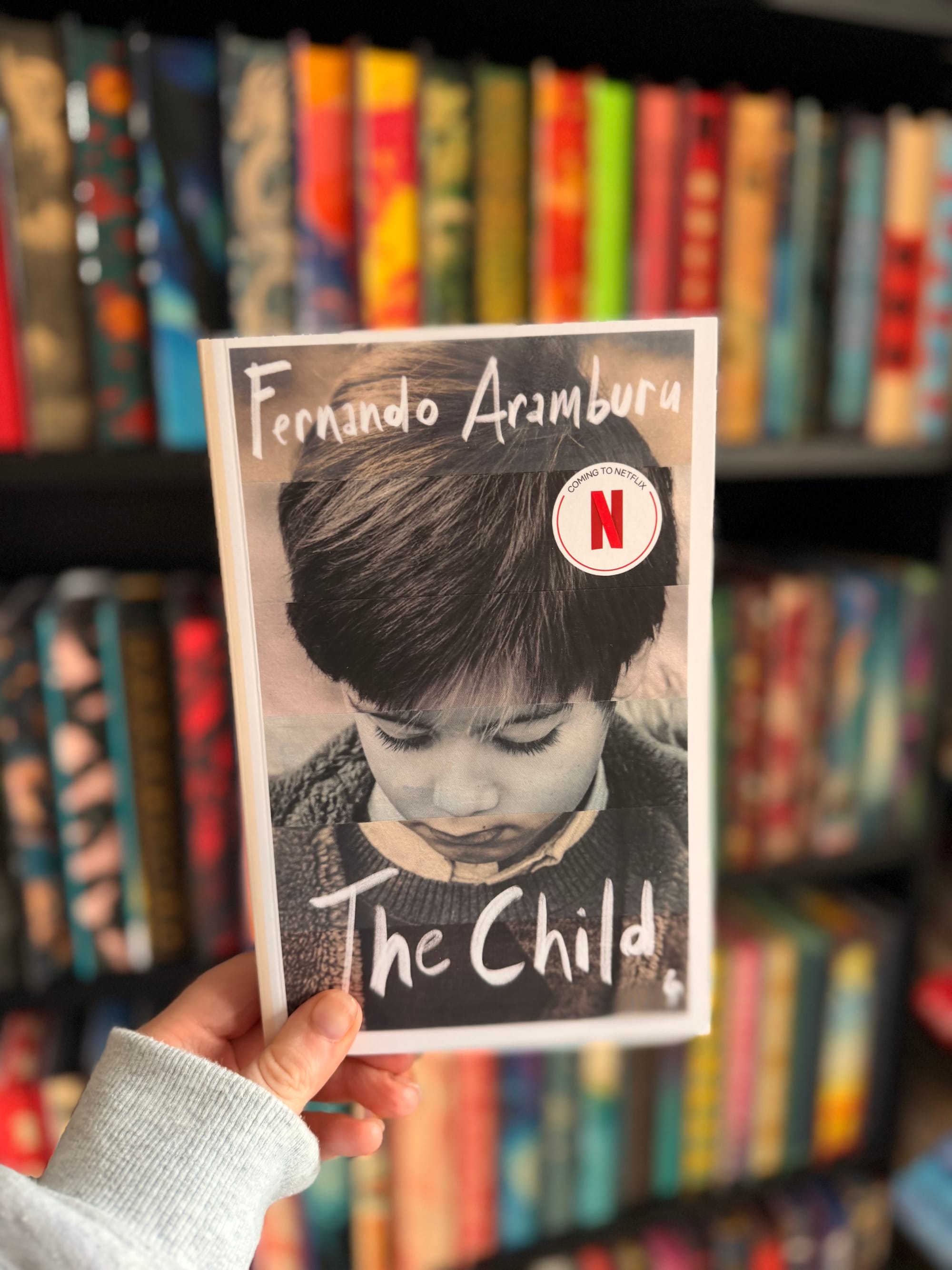 A hand holding a copy of The Child by Fernando Aramburu in front of a colourful, softly blurred bookshelf filled with books.