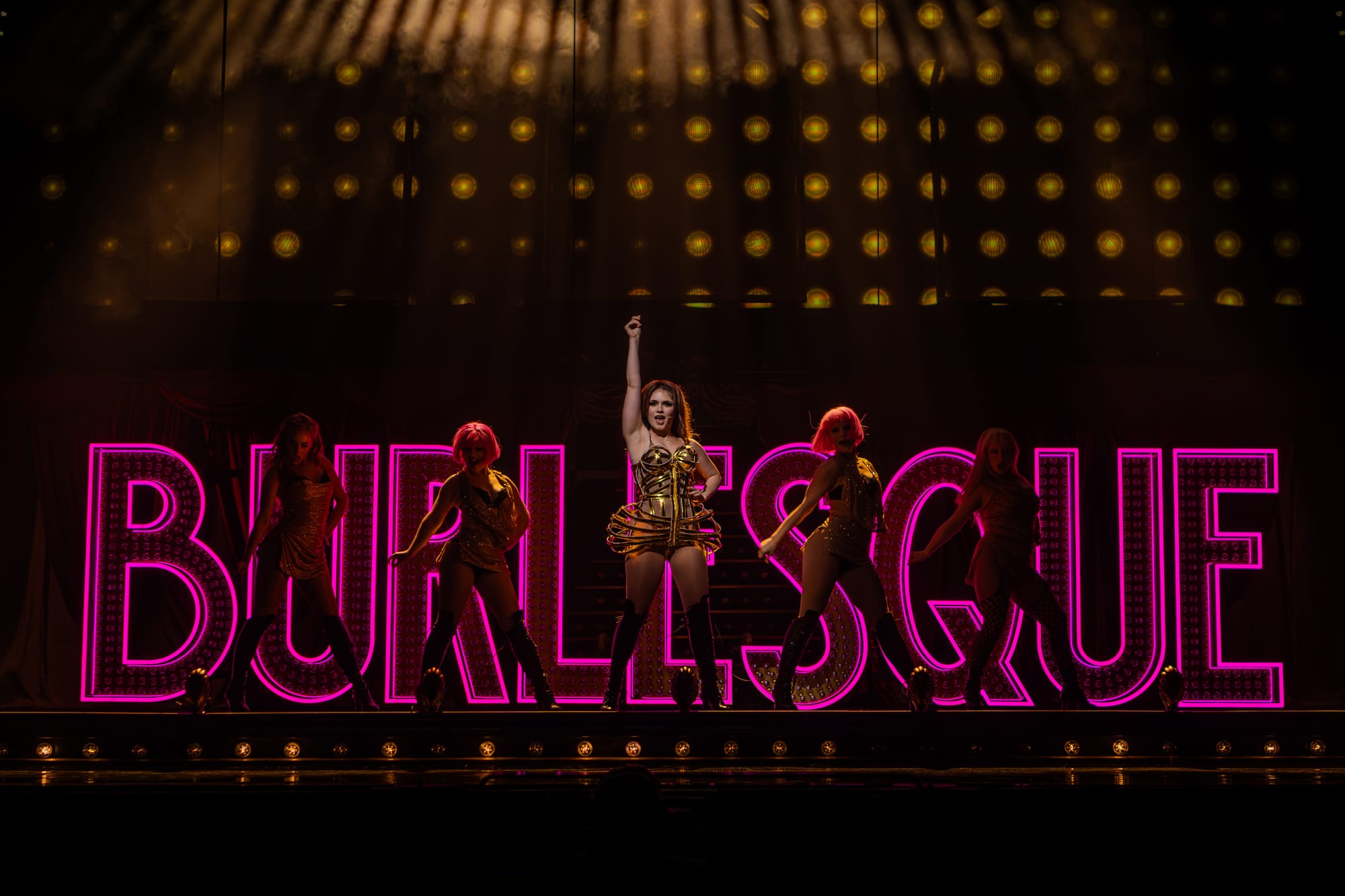 Performer in a gold costume centre stage with four dancers in front of a large pink neon ‘BURLESQUE’ sign, under dramatic golden theatre lighting.