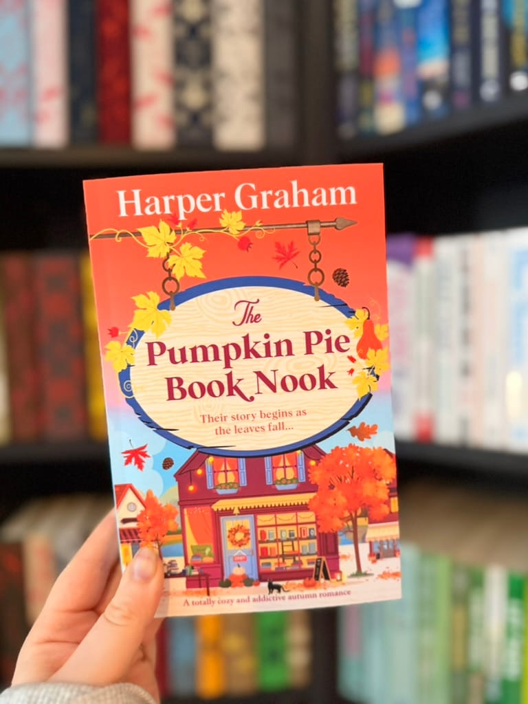 A hand holds a copy of The Pumpkin Pie Book Nook by Harper Graham in front of blurred bookshelves. The colourful autumn-themed cover features orange leaves, pumpkins and a cosy bookshop illustration.