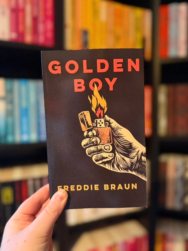 A hand holds a copy of Golden Boy by Freddie Braun in front of colour-coordinated bookshelves, with the book’s dark cover and illustrated flaming lighter in sharp focus.