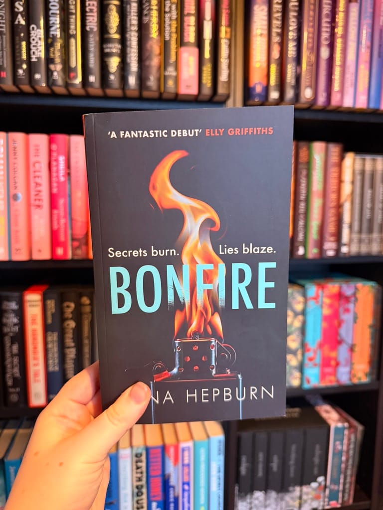 A hand holds a copy of Bonfire by Anna Hepburn in front of colour-coordinated bookshelves, with the dark cover and flaming lighter design in sharp focus.