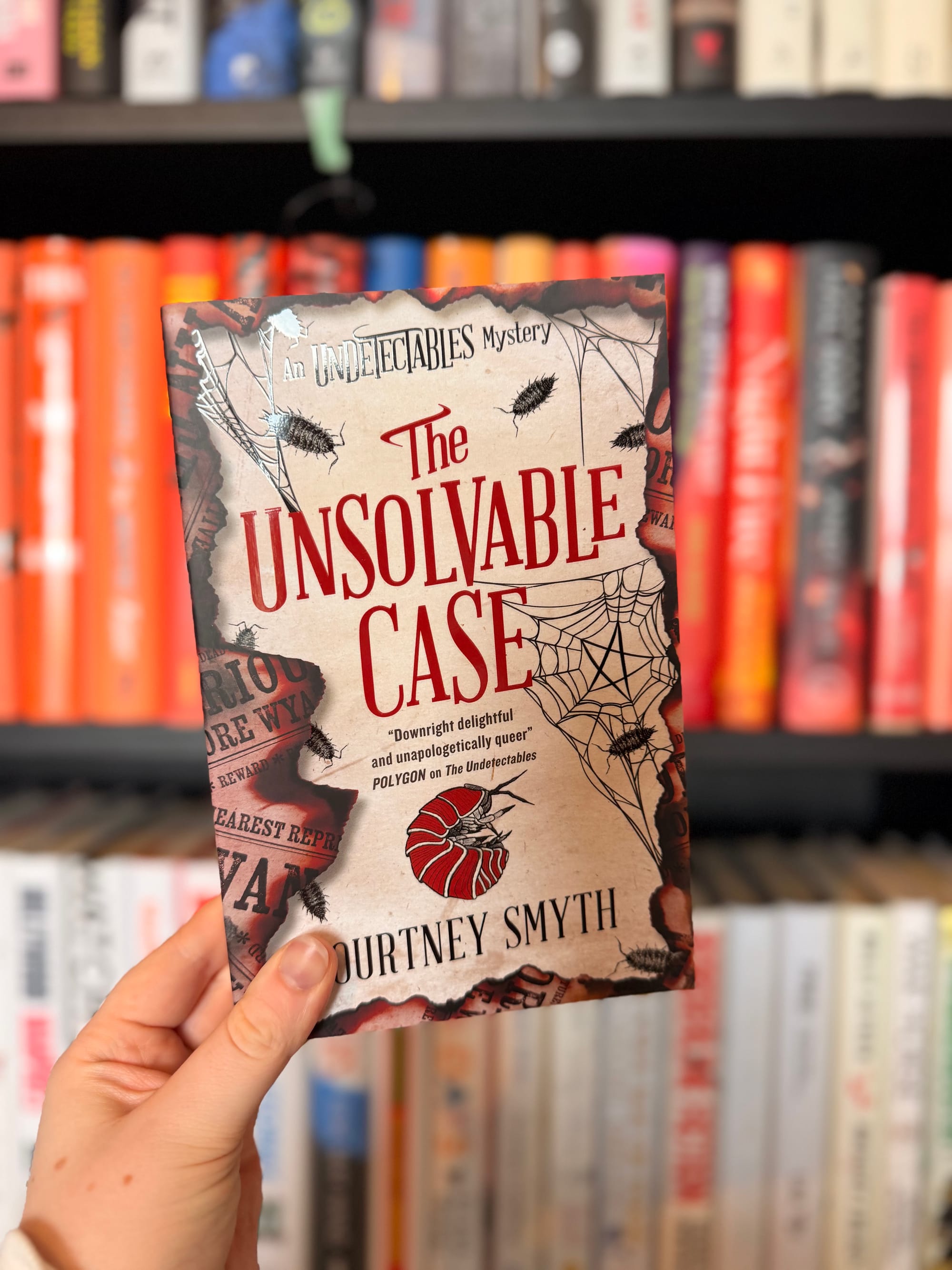 A hand holds a copy of The Unsolvable Case by Courtney Smyth in front of blurred bookshelves. The cream-coloured cover features red lettering, cobwebs, insects and torn newspaper-style details.