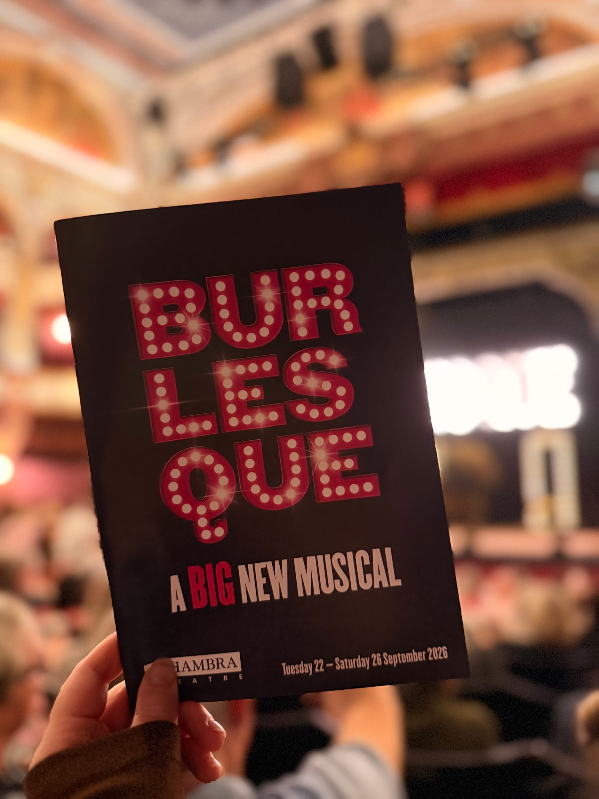 Burlesque theatre programme held up inside the Alhambra Theatre, with the stage softly blurred in the background.