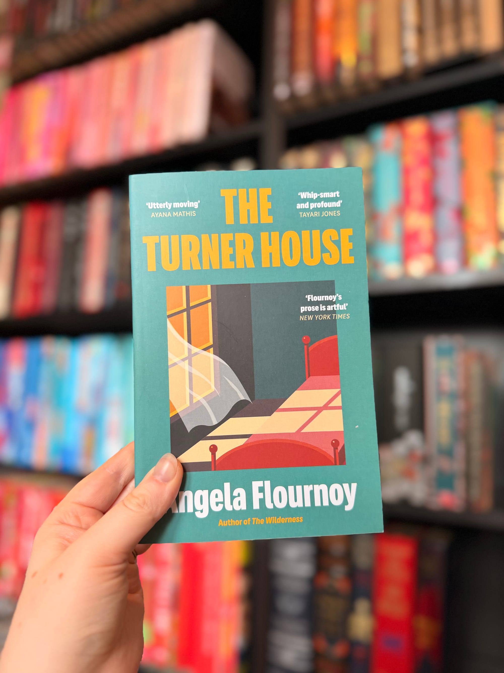 A hand holding a copy of The Turner House by Angela Flournoy in front of colourful, blurred bookshelves. The book has a teal cover with bold yellow lettering and an illustrated bedroom scene.