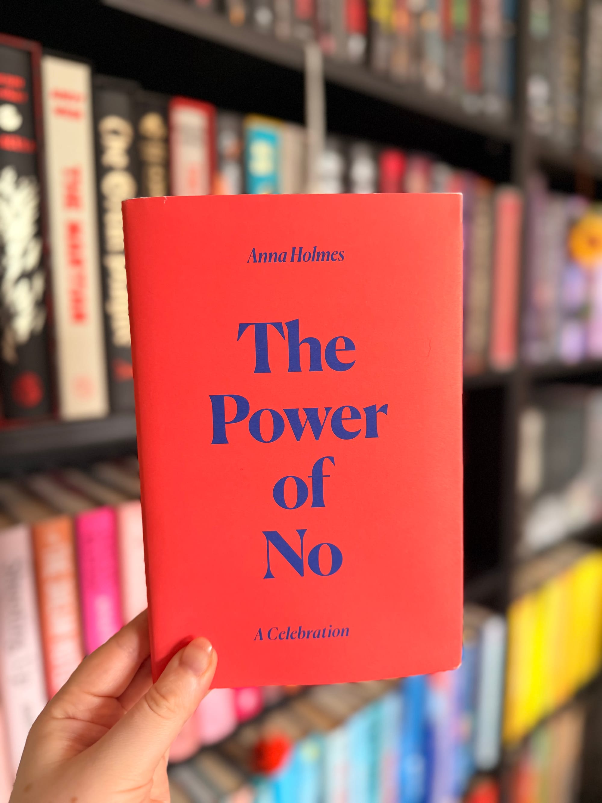 A hand holding a red copy of The Power of No: A Celebration by Anna Holmes in front of colourful, blurred bookshelves.