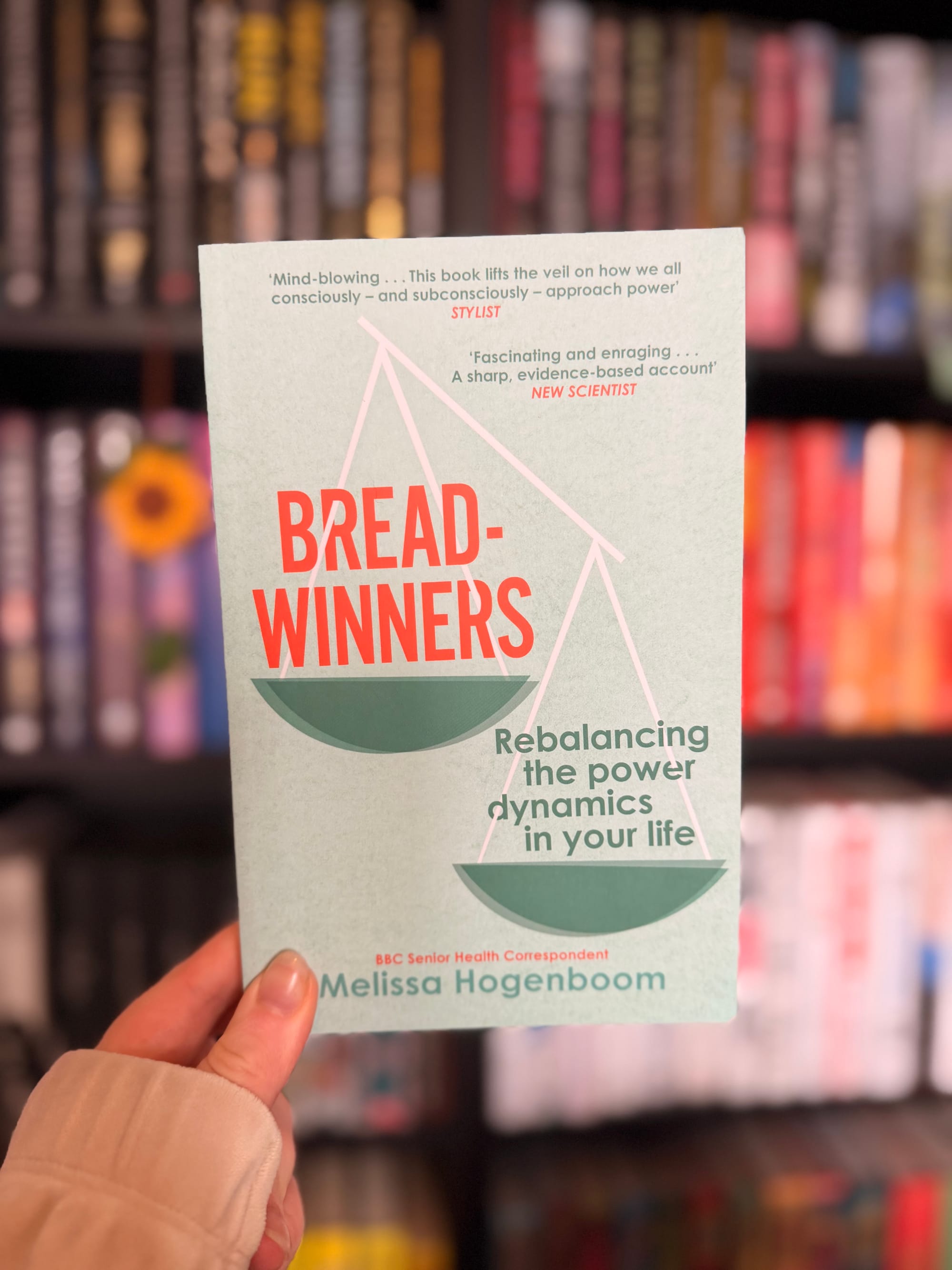 A hand holding Breadwinners: Rebalancing the Power Dynamics in Your Life by Melissa Hogenboom, with blurred bookshelves in the background.