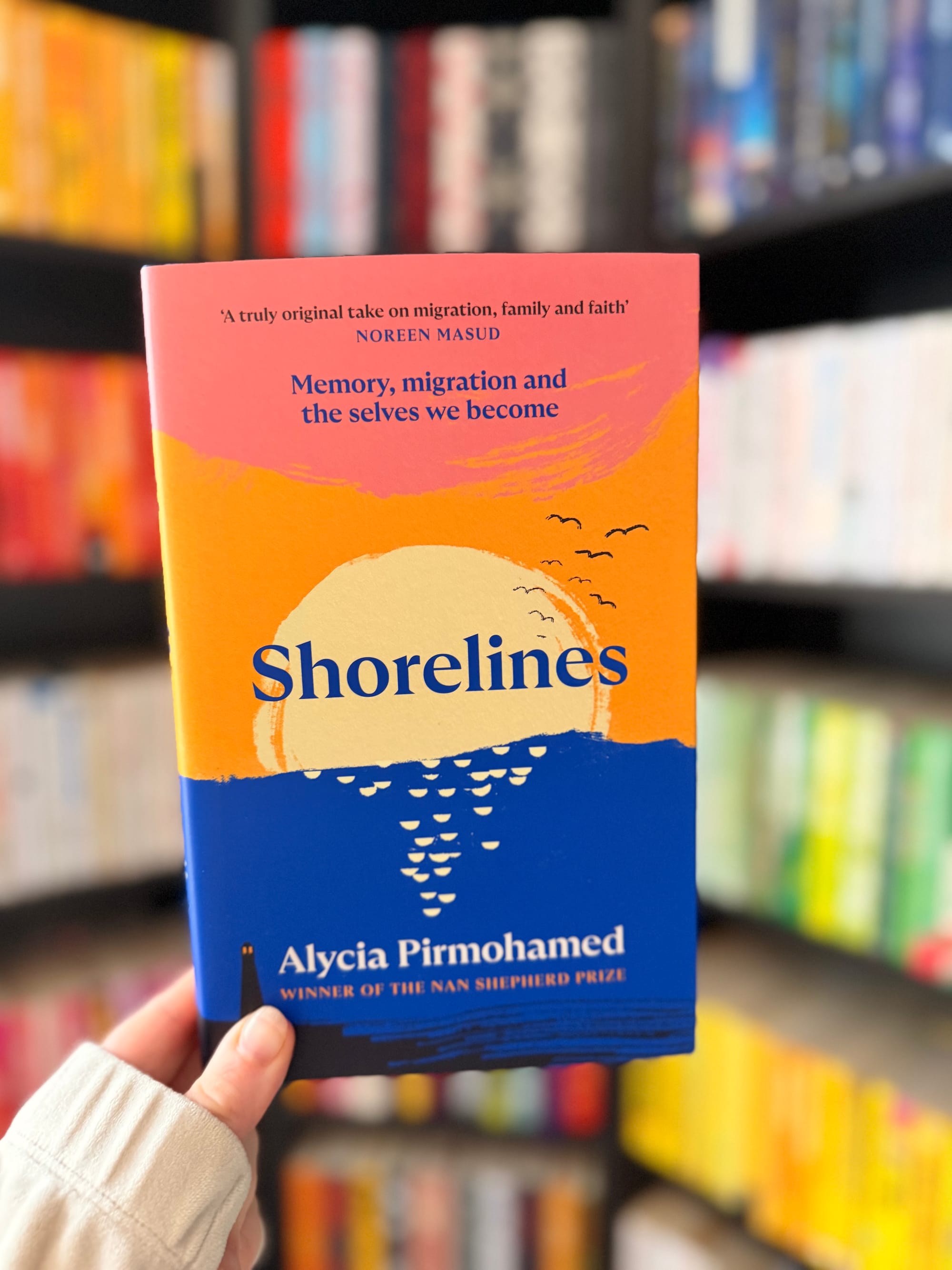 A hand holding Shorelines by Alycia Pirmohamed, featuring a bright orange, pink and blue cover, in front of colour-coordinated bookshelves.