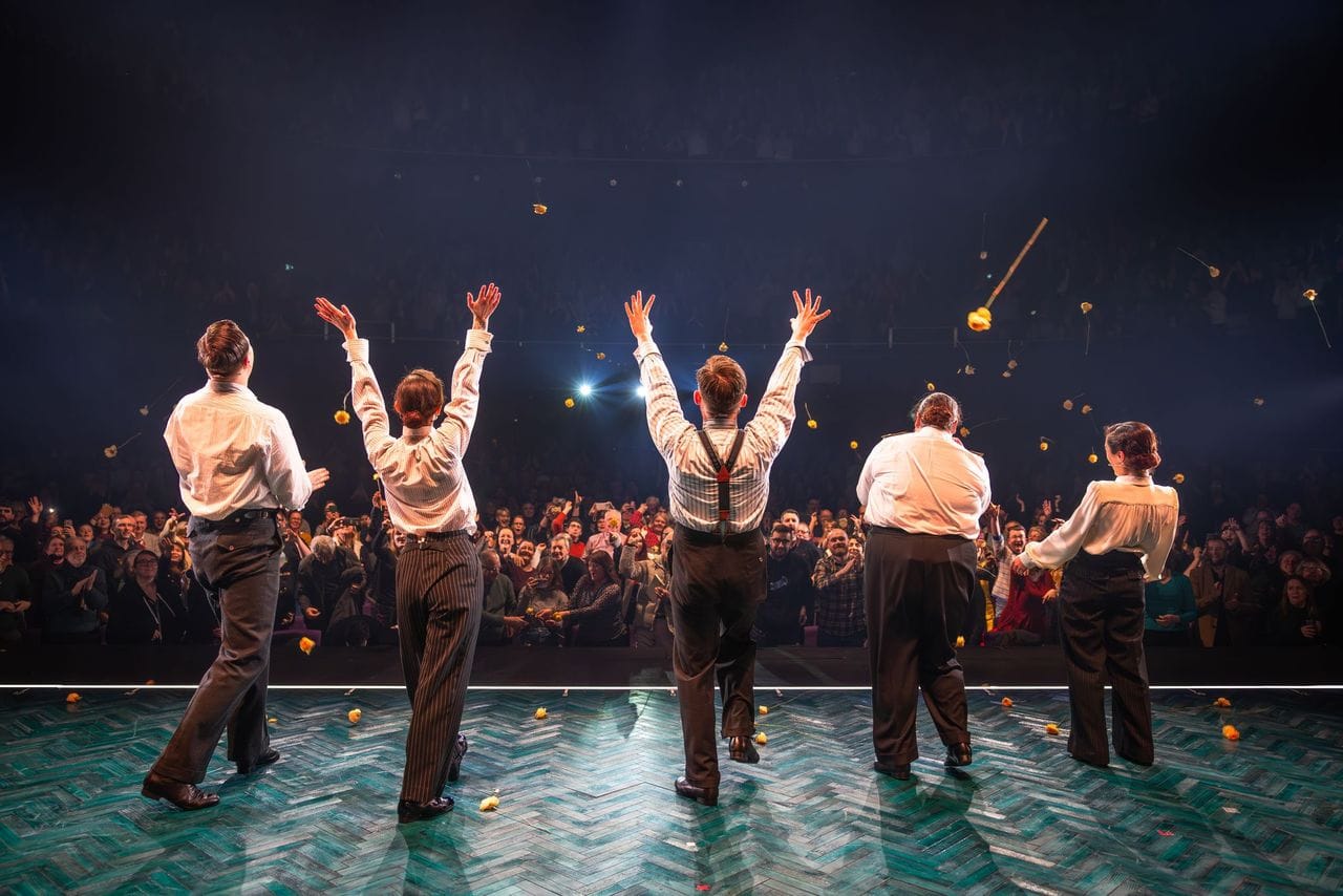 Five cast members take their bows on stage in front of a cheering audience.