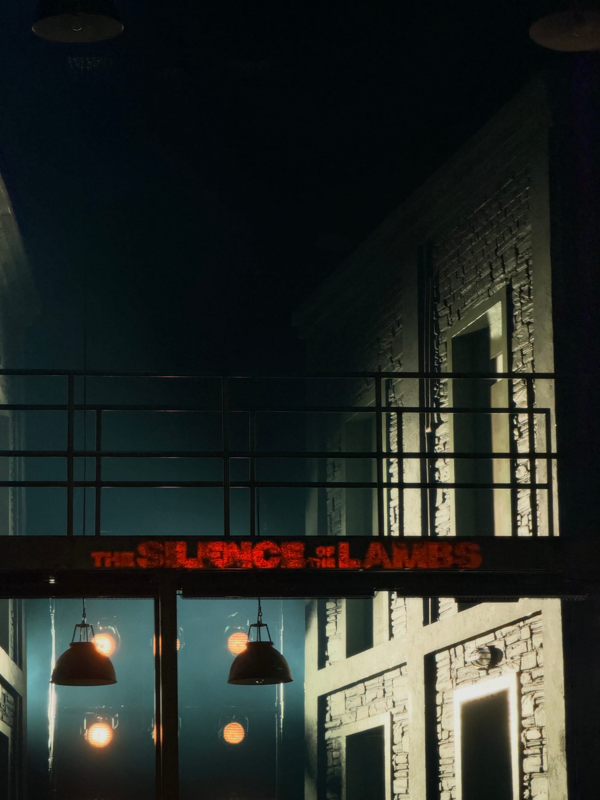 A dark, atmospheric stage set from The Silence of the Lambs, featuring an industrial-style upper walkway with black metal railings, exposed brick walls, illuminated doorways and hanging lights. The production title glows red across the centre of the set.