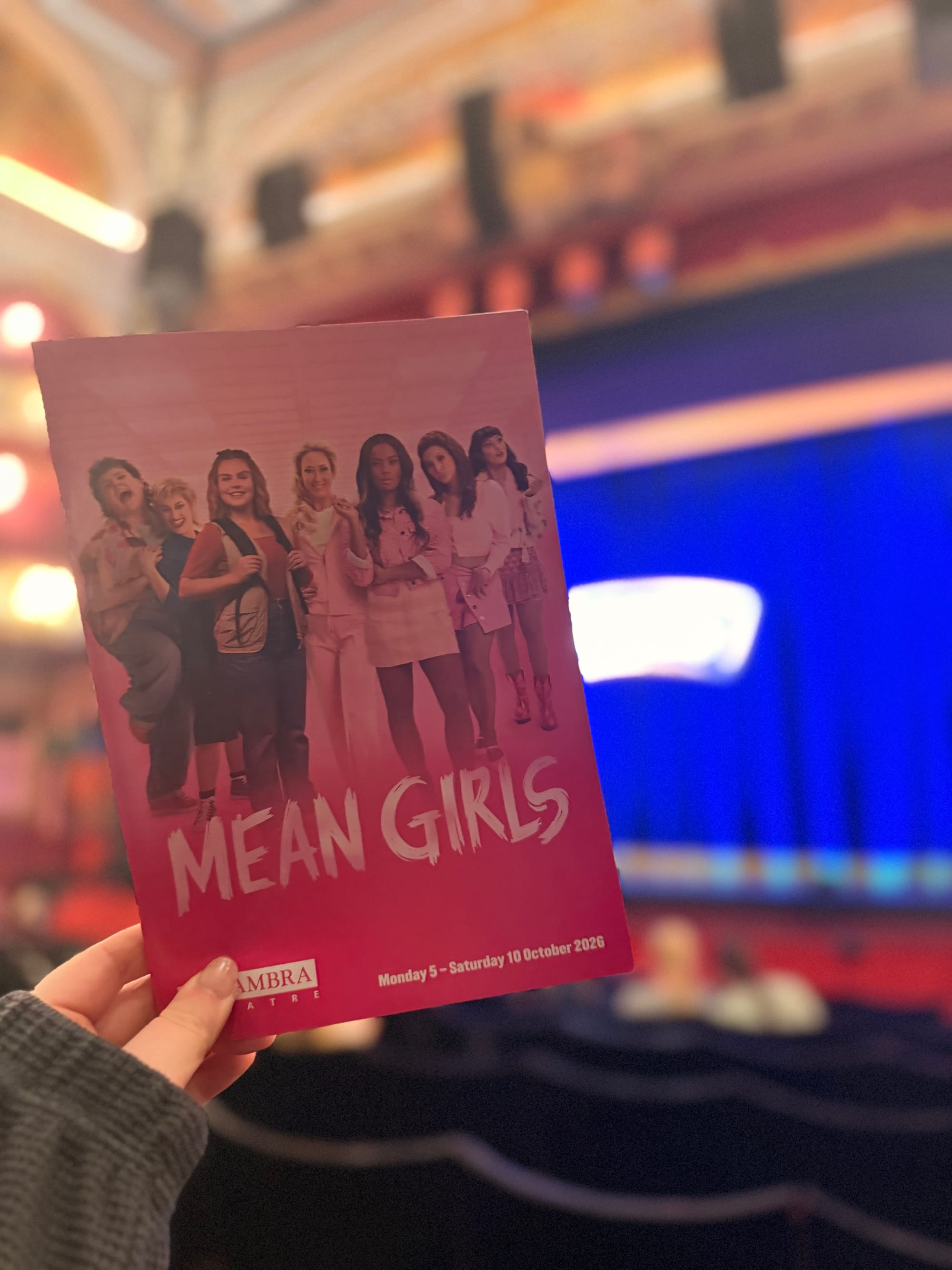 A hand holding a Mean Girls theatre programme inside the venue, with the pink programme cover in focus and the stage blurred in the background.