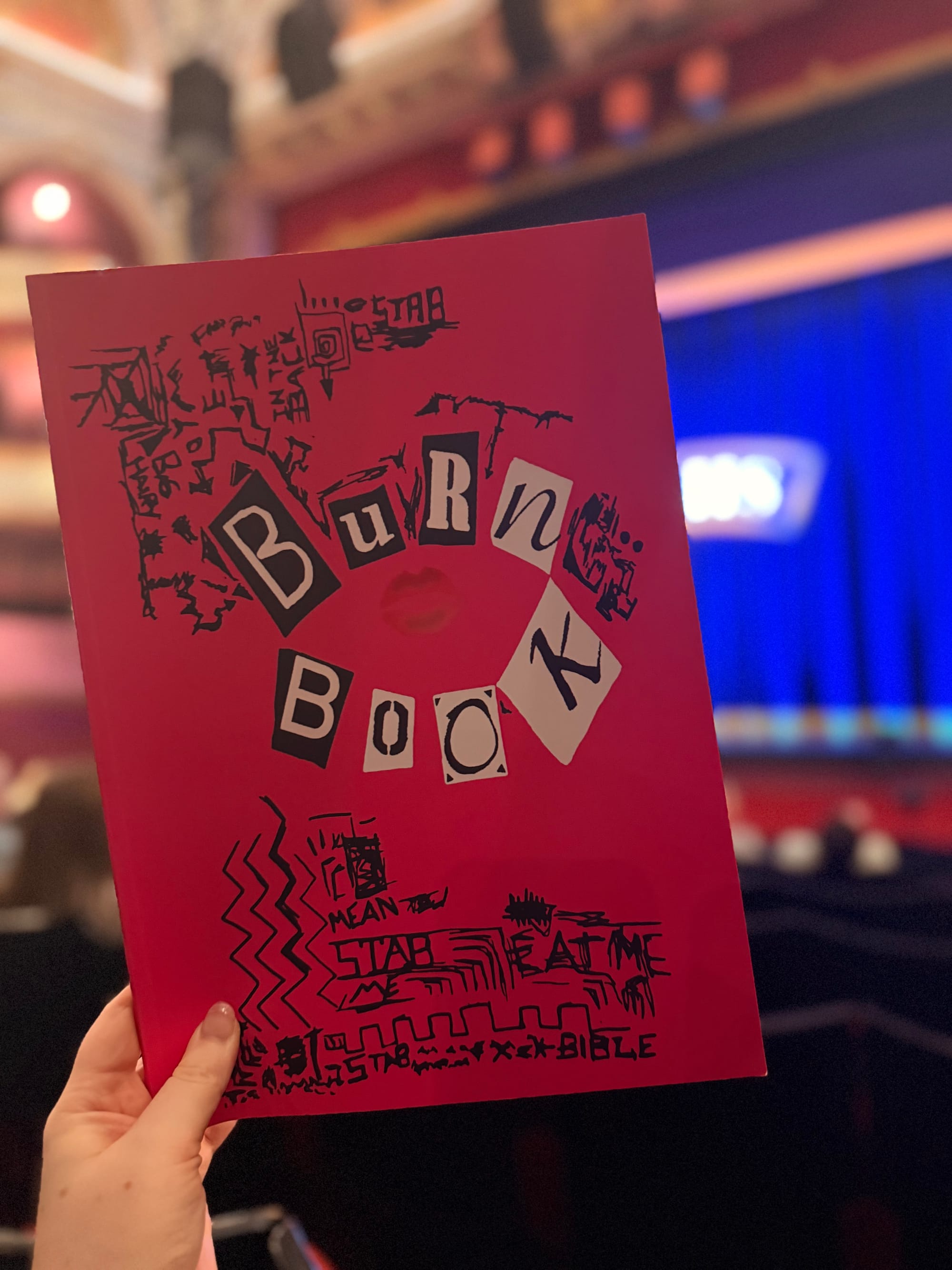 A hand holding a bright pink Mean Girls “Burn Book” programme featuring ransom-note style lettering and black doodles, with the theatre stage blurred behind it.