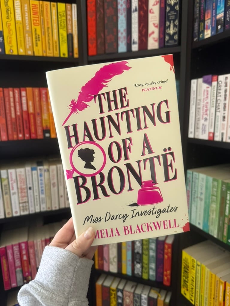 Hand holds The Haunting of a Brontë by Amelia Blackwell in front of colorful bookshelves.