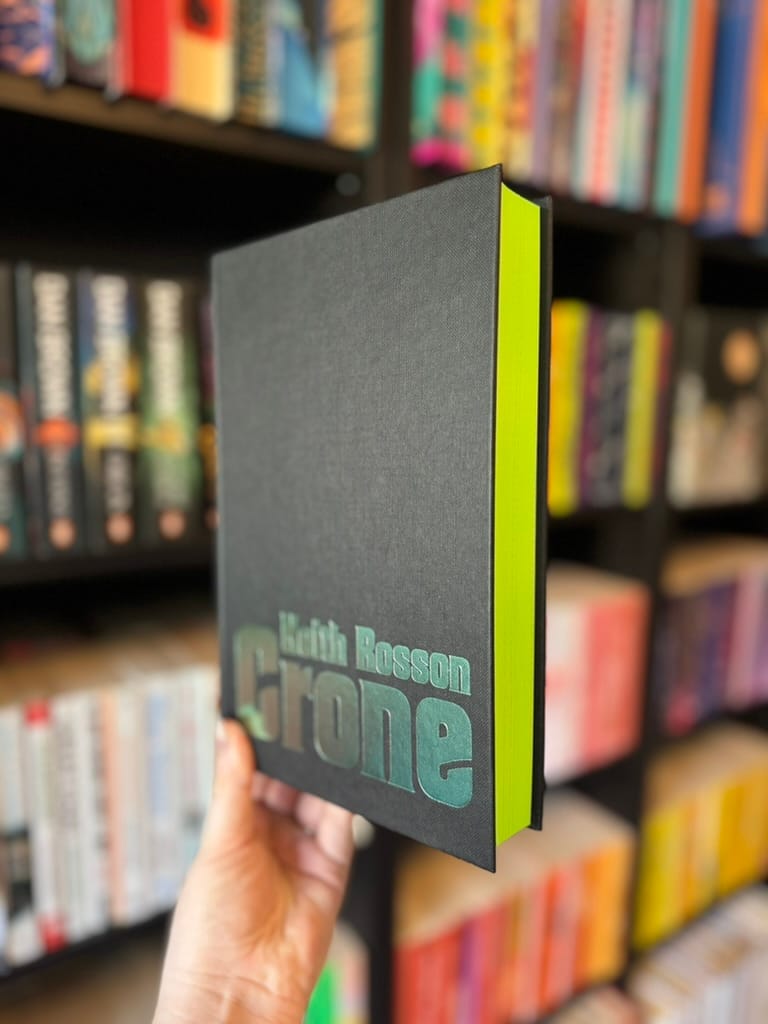 Hand holding a book titled "Keith Rosson Crone" with a black cover and neon yellow pages, against a colorful bookshelf background.