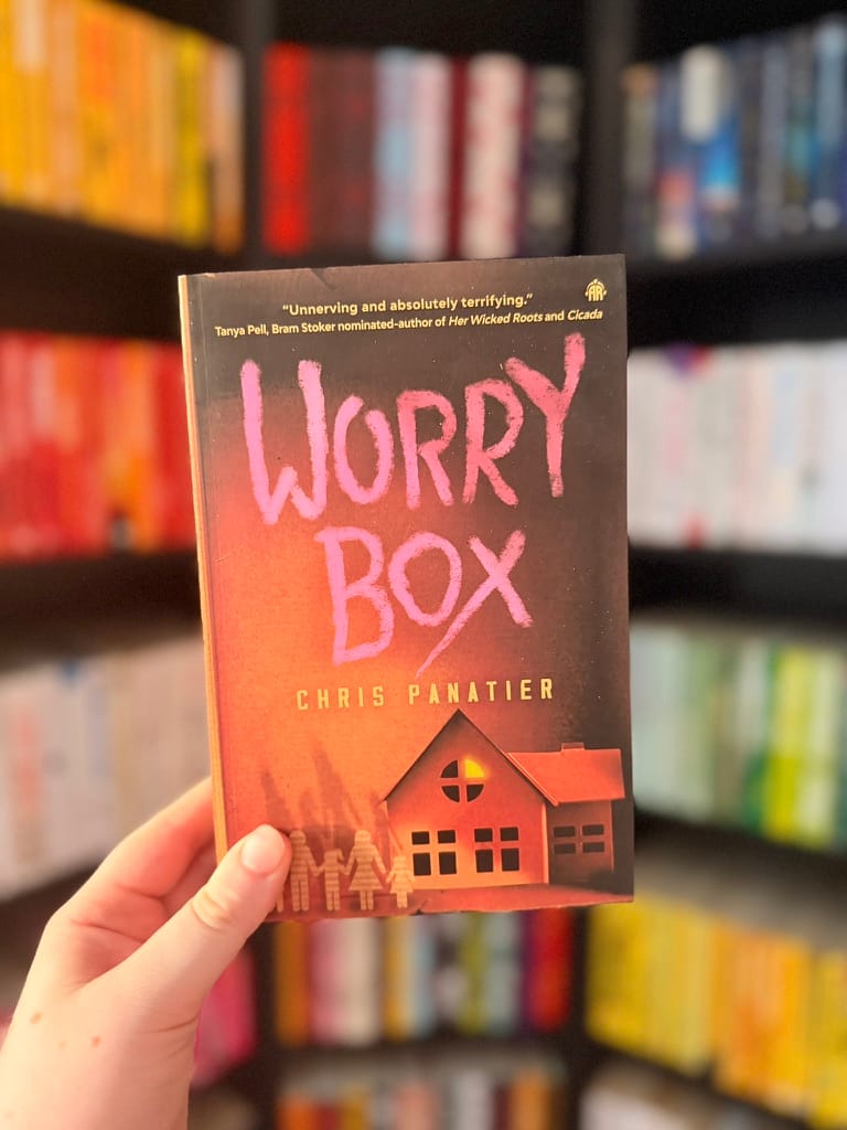 A hand holds a copy of Worry Box by Chris Panatier in front of a blurred bookshelf filled with colourful books. 