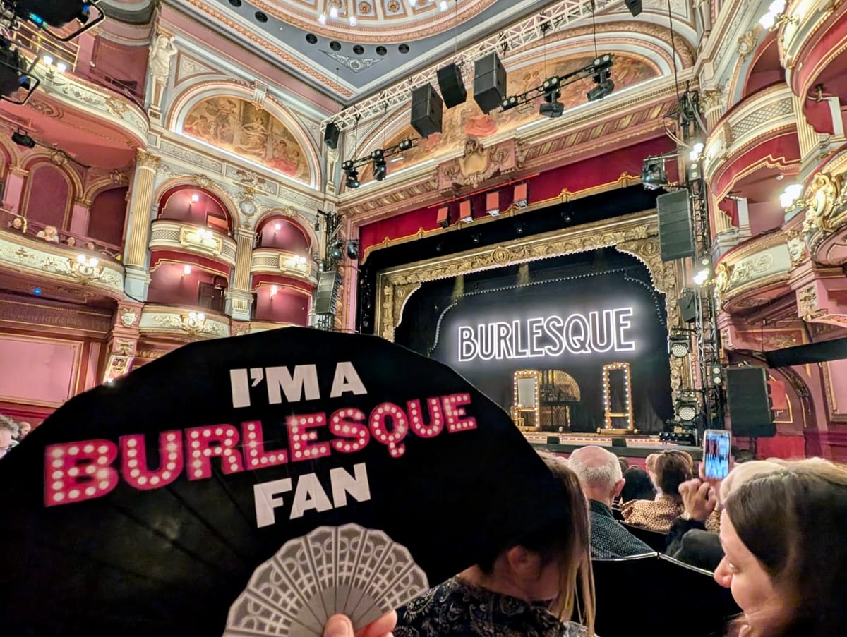 Interior of the Alhambra Theatre with the Burlesque stage in the background and a black fan in the foreground reading “I’m a Burlesque Fan"