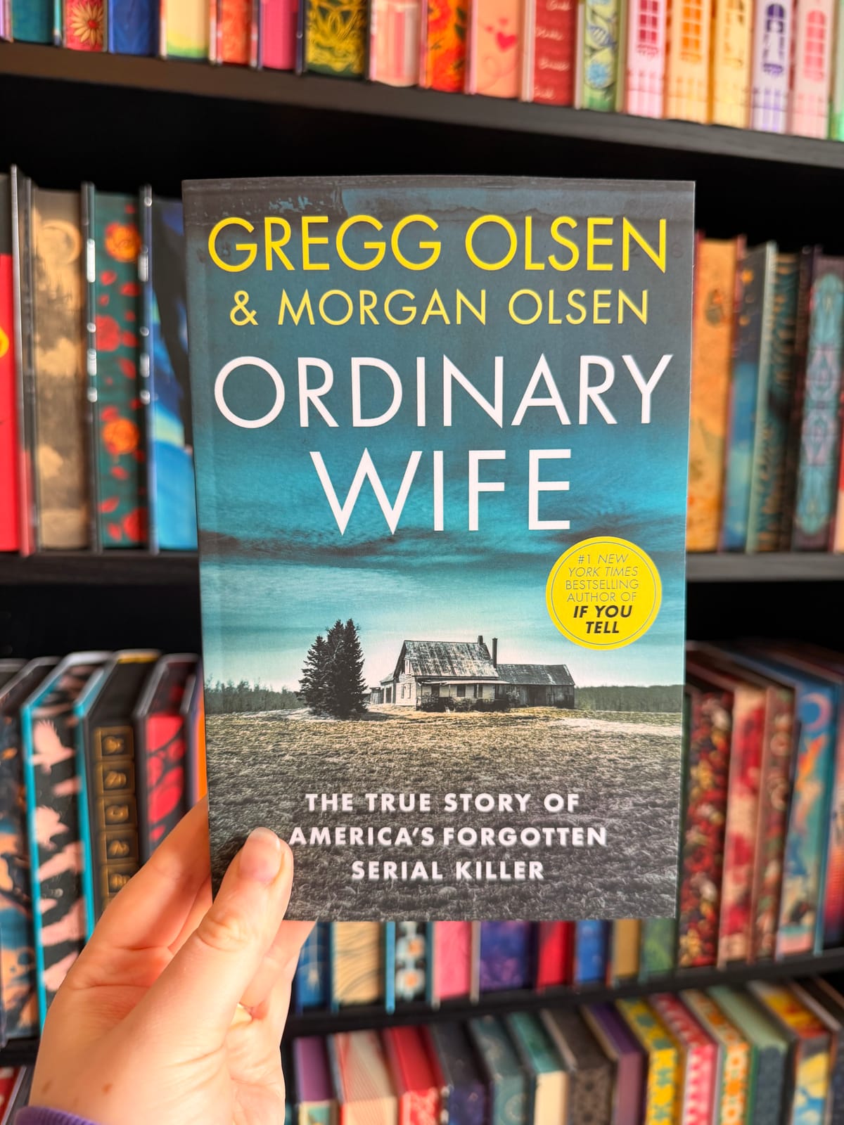 A hand holds a copy of Ordinary Wife by Gregg Olsen and Morgan Olsen in front of colourful bookshelves. The cover features a remote farmhouse beneath a dark blue sky.