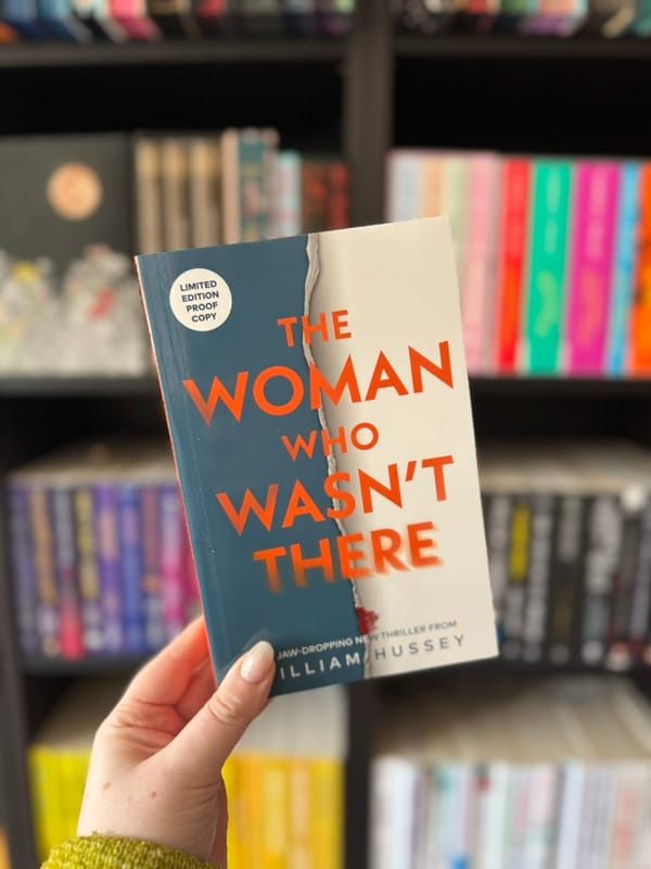 A hand holds a book titled "The Woman Who Wasn't There" by William Hussey. Background shows colorful bookshelves, creating a vibrant scene.