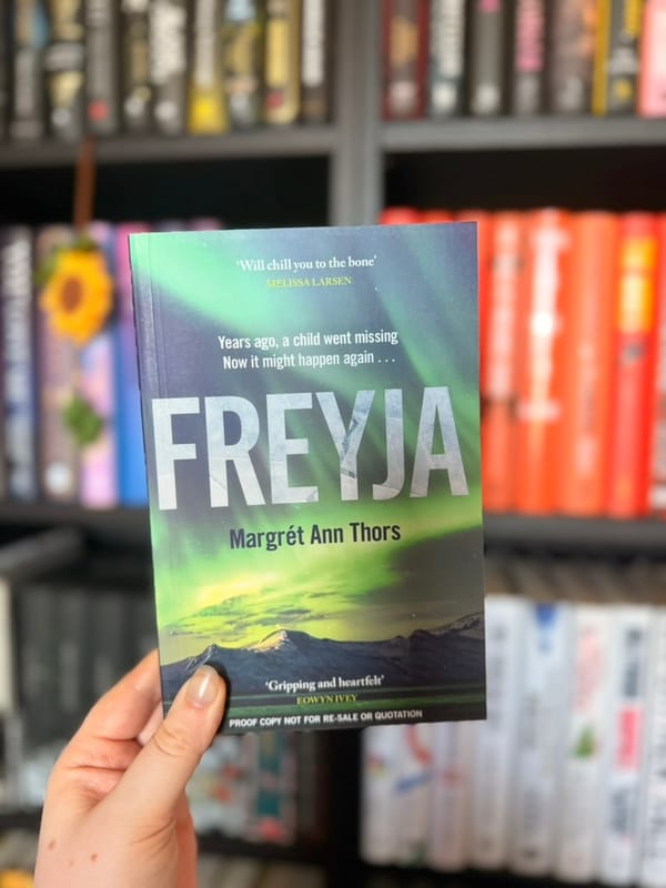 A hand holds the proof copy of Freyja by Margrét Ann Thors in front of softly blurred bookshelves.