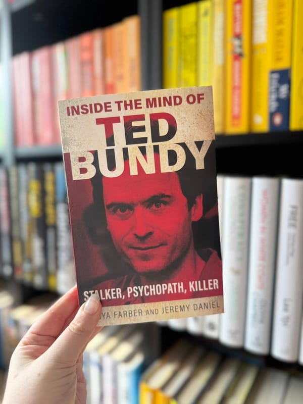 Hand holding a book titled Inside the Mind of Ted Bundy with a red-toned cover, in front of a colorful bookshelf background.